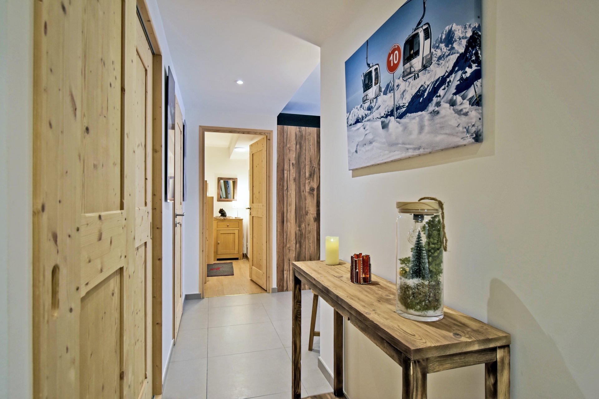 Couloir intérieur avec meubles en bois, tableau de montagne avec télécabines, décor de Noël dans un bocal, porte en bois, sol en carrelage, éclairage encastré.