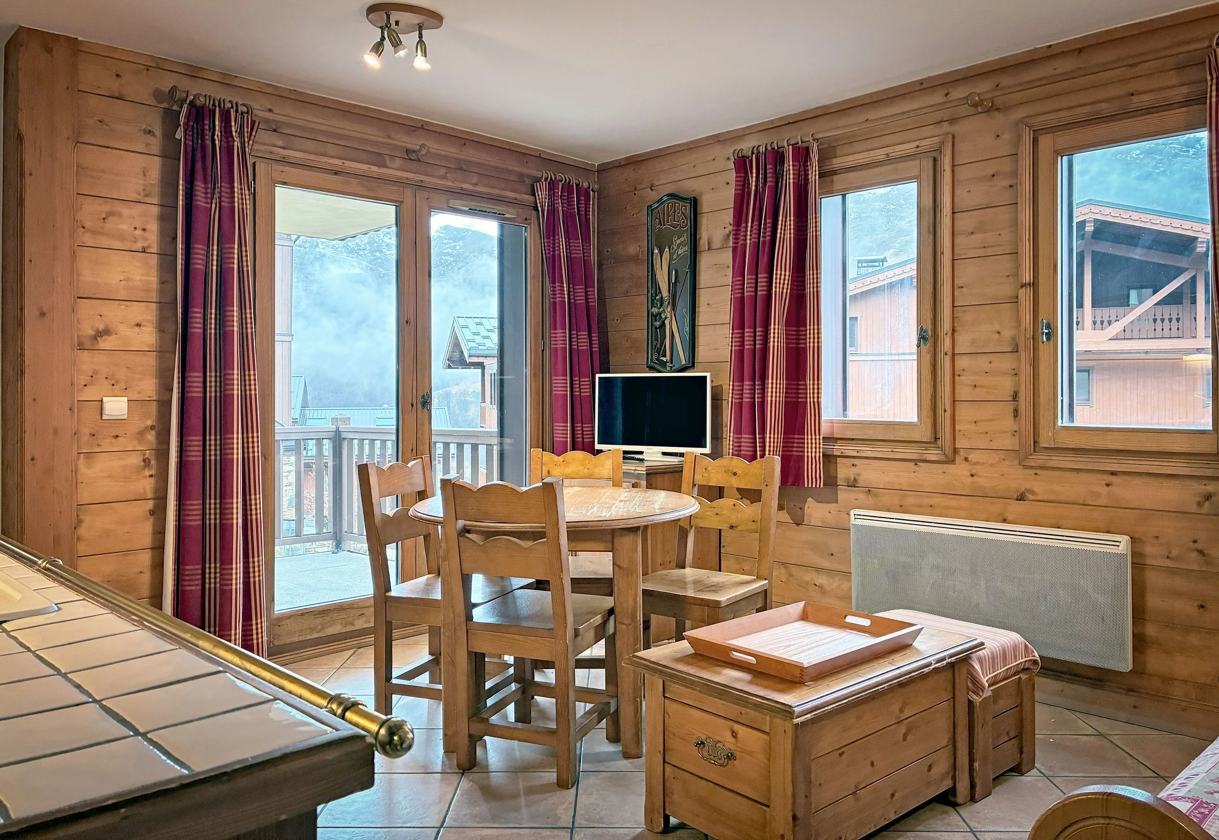 Intérieur en bois avec table ronde, chaises, télévision, fenêtres avec rideaux rouges, porte-fenêtre donnant sur un balcon, murs en bois, chauffage électrique.