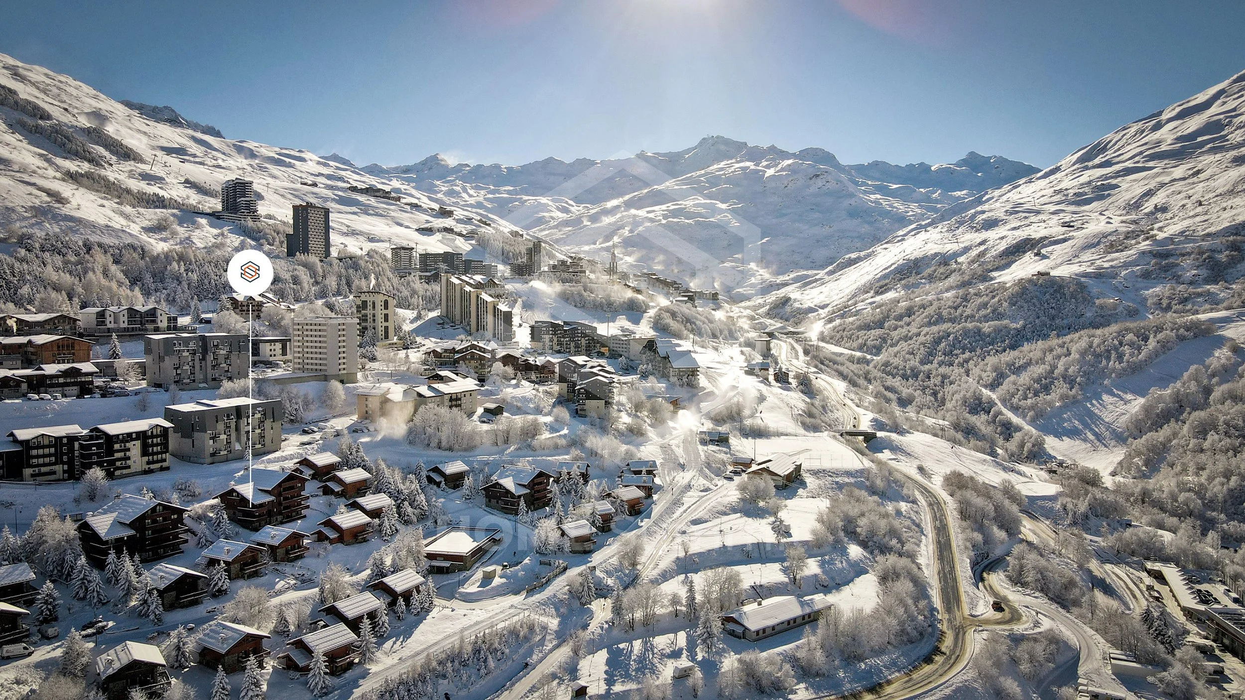 Vue aérienne d'un village alpin enneigé avec des bâtiments modernes et chalets en bois, entouré de montagnes enneigées sous un ciel clair.