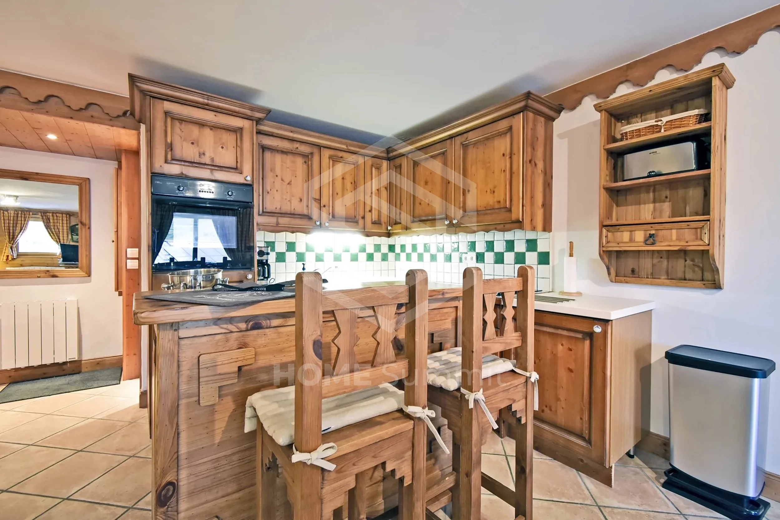 Cuisine en bois avec comptoir, chaises en bois avec coussins, dosseret en carreaux verts et blancs, four encastré, étagère en bois, poubelle à compost.