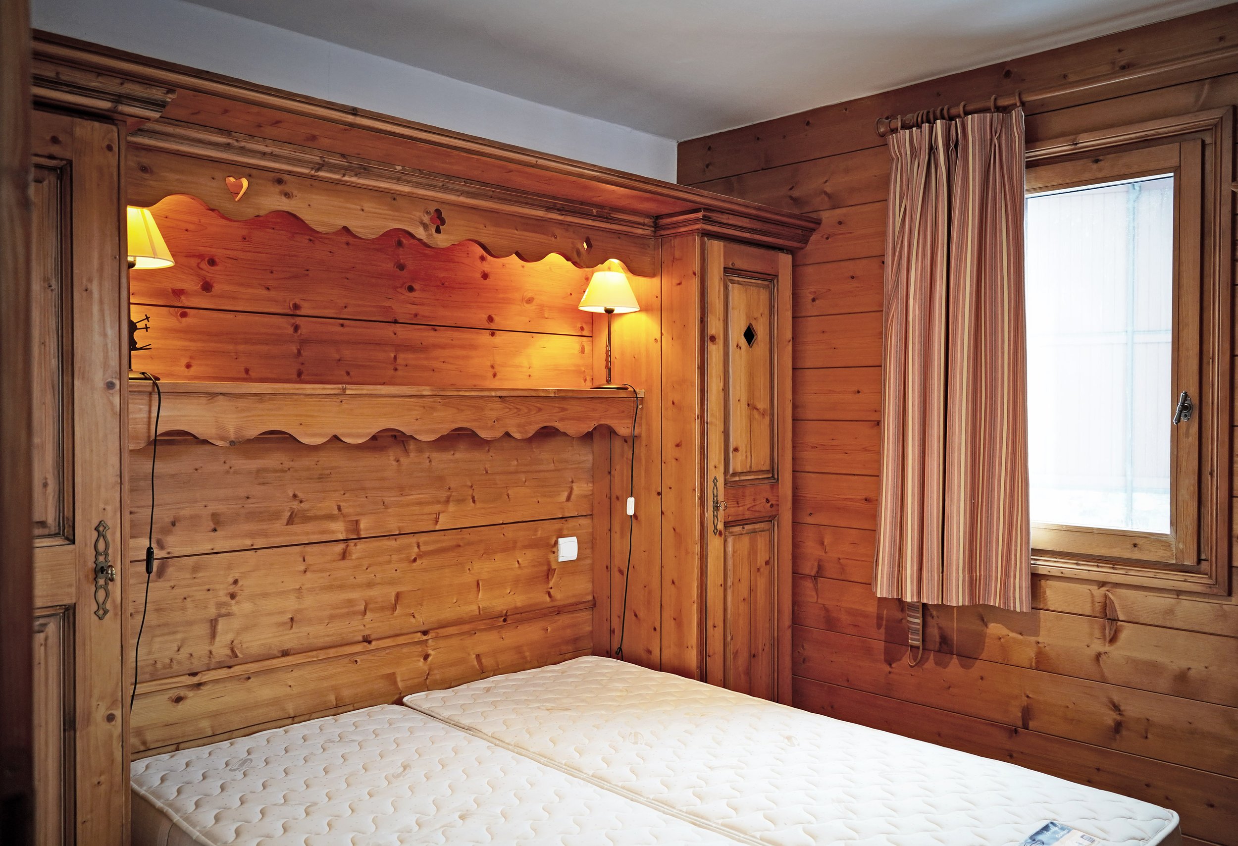 Chambre en bois avec un lit sans draps, une fenêtre avec rideau rayé, et deux lampes murales encadrant la tête du lit.