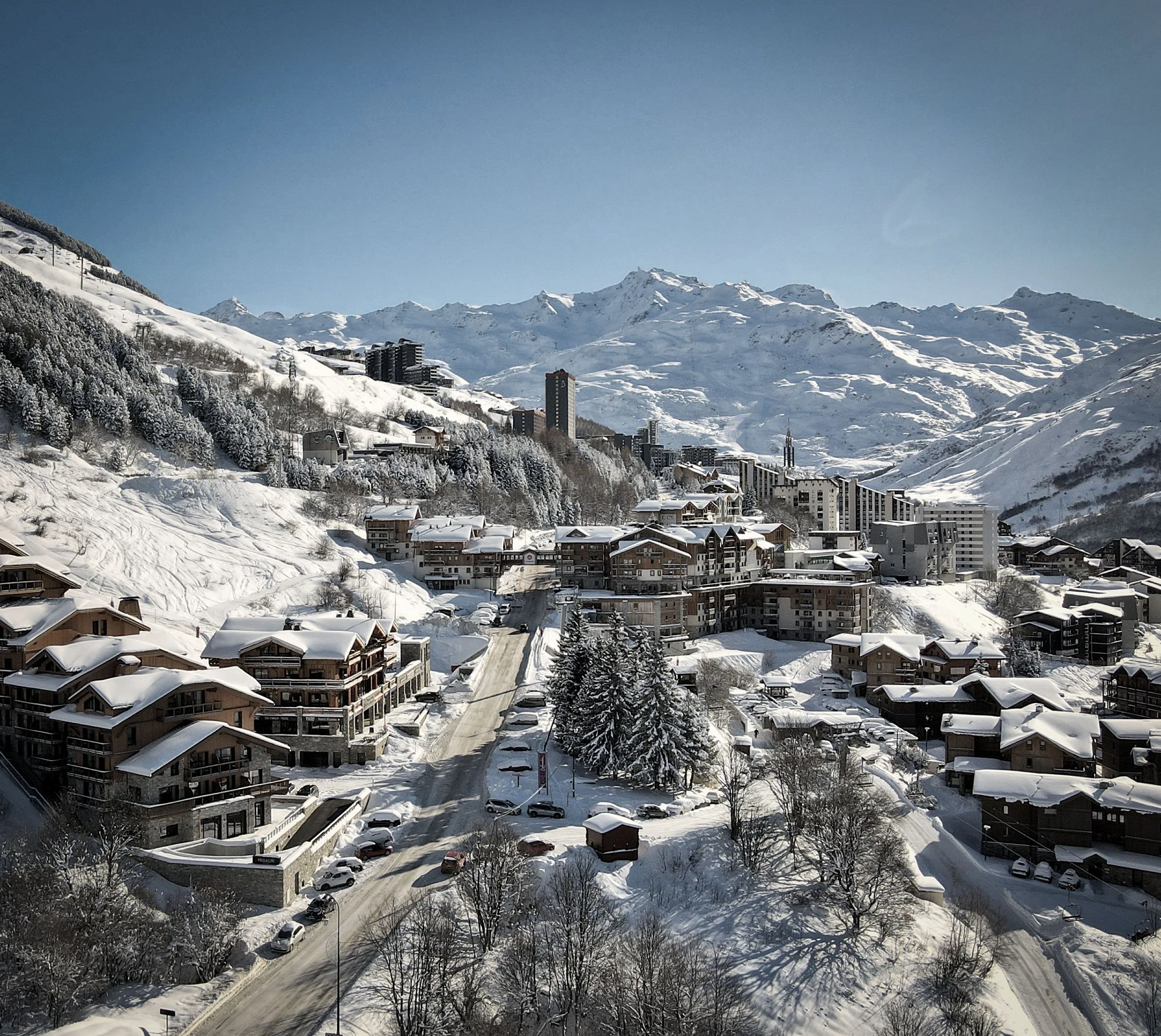 Village alpin enneigé avec des bâtiments, des routes, des arbres et des montagnes en arrière-plan sous un ciel clair.