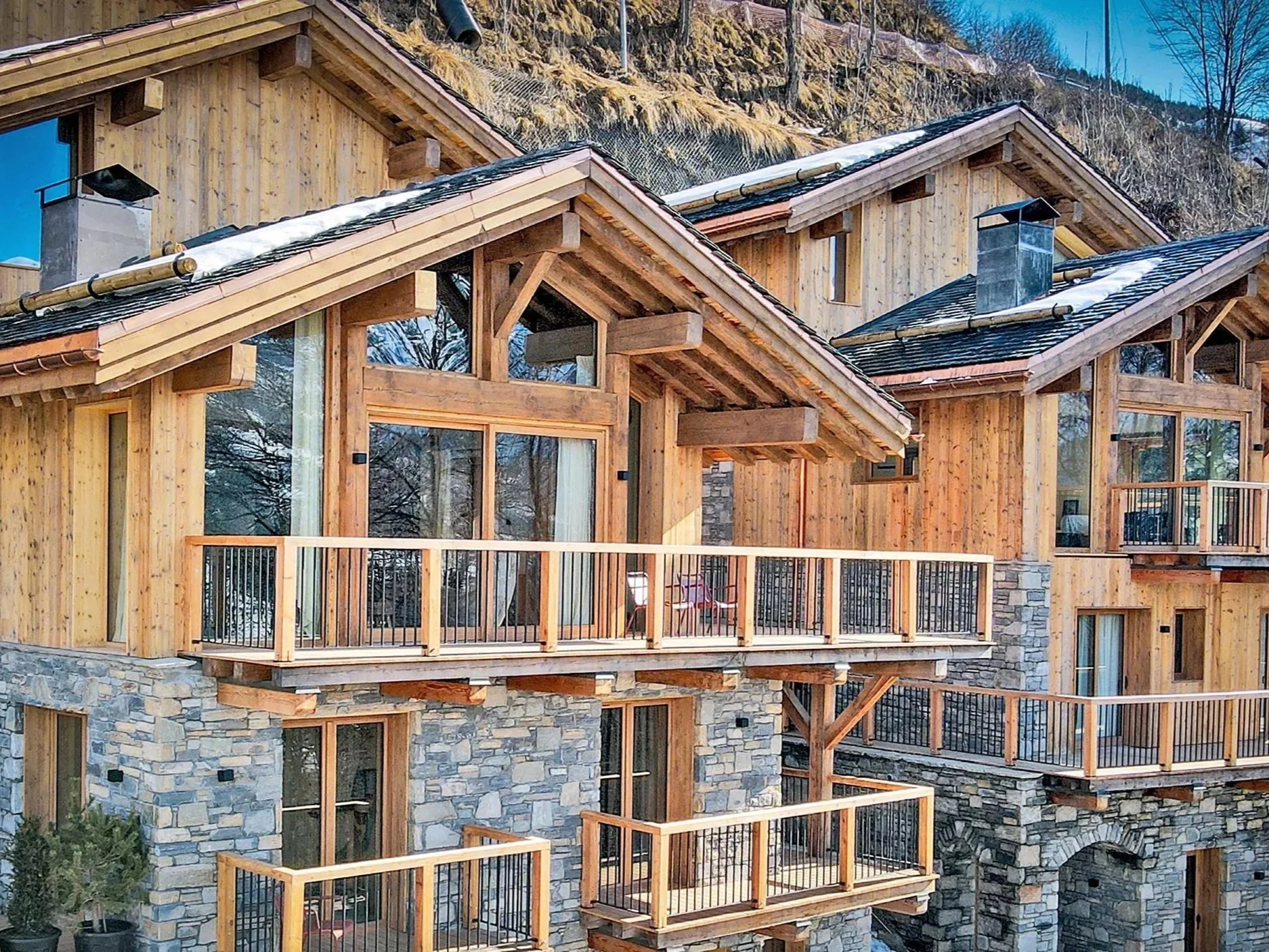 Façade d’un chalet de prestige à proximité de Saint-Martin-de-Belleville, associant pierre naturelle, bardage en bois, grandes baies vitrées, balcons et terrasses dans un environnement alpin.