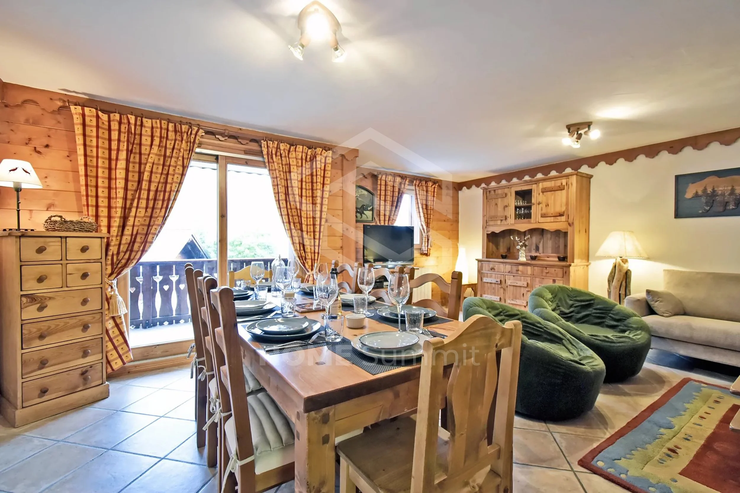 Salon rustique avec meubles en bois, table à manger prête pour un repas, fauteuils verts, canapé beige, armoires en bois, fenêtres avec rideaux à carreaux, télévision, éclairage chaleureux.