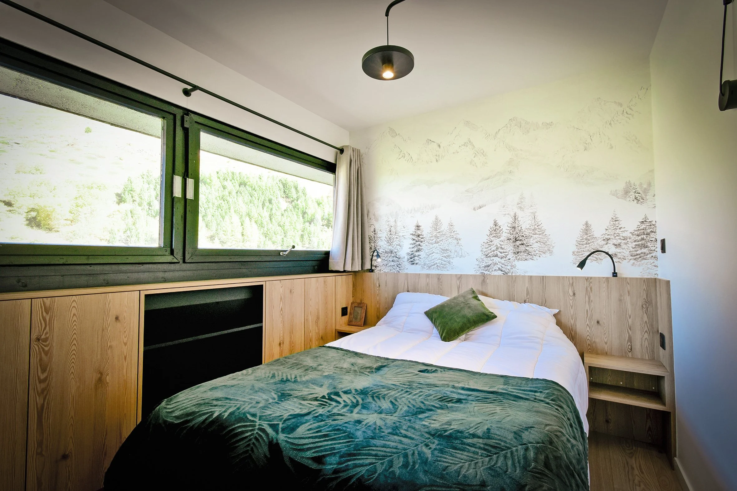 Chambre avec lit double, mur avec décor de forêt et montagnes, grande fenêtre laissant entrer la lumière naturelle, mobilier en bois clair, lampe de chevet