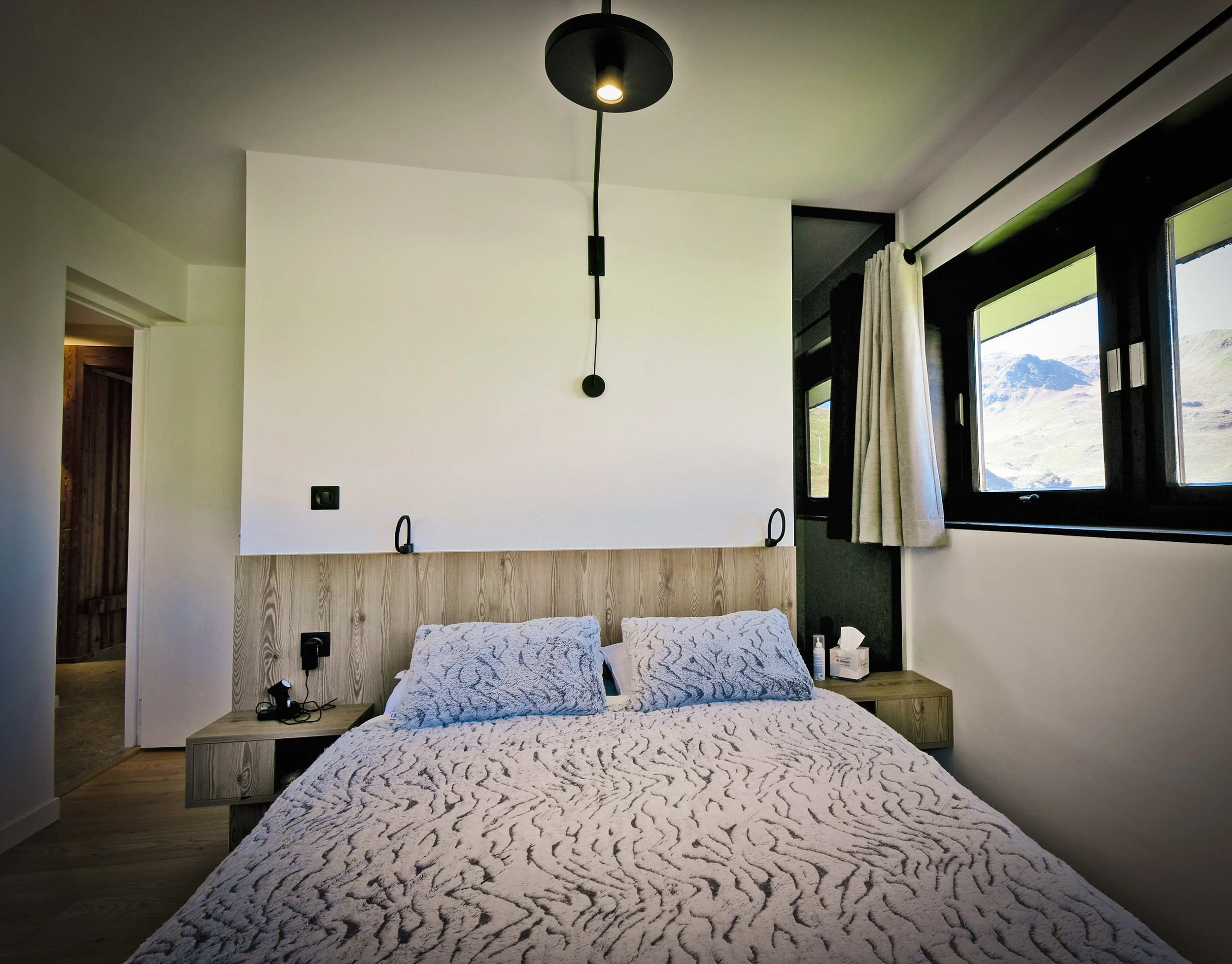 Chambre moderne avec lit, deux oreillers, tête de lit en bois, lampes murales, fenêtres avec vue sur montagnes, rideaux blancs et noirs, mobilier en bois clair.