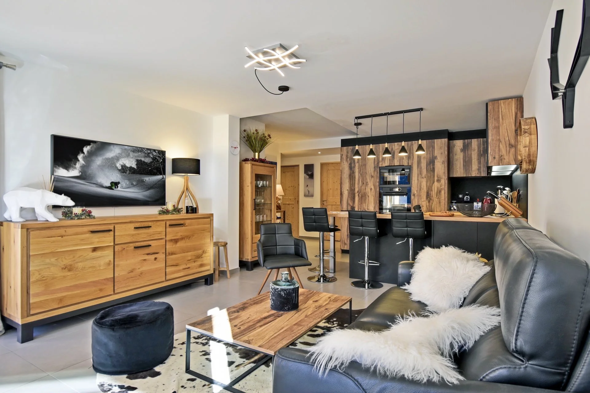 Salon et cuisine modernes avec mobilier en bois naturel et noir, canapé en cuir noir avec coussins en fourrure blanche, télévision, table basse en bois, cuisine équipée avec bar et chaises hautes