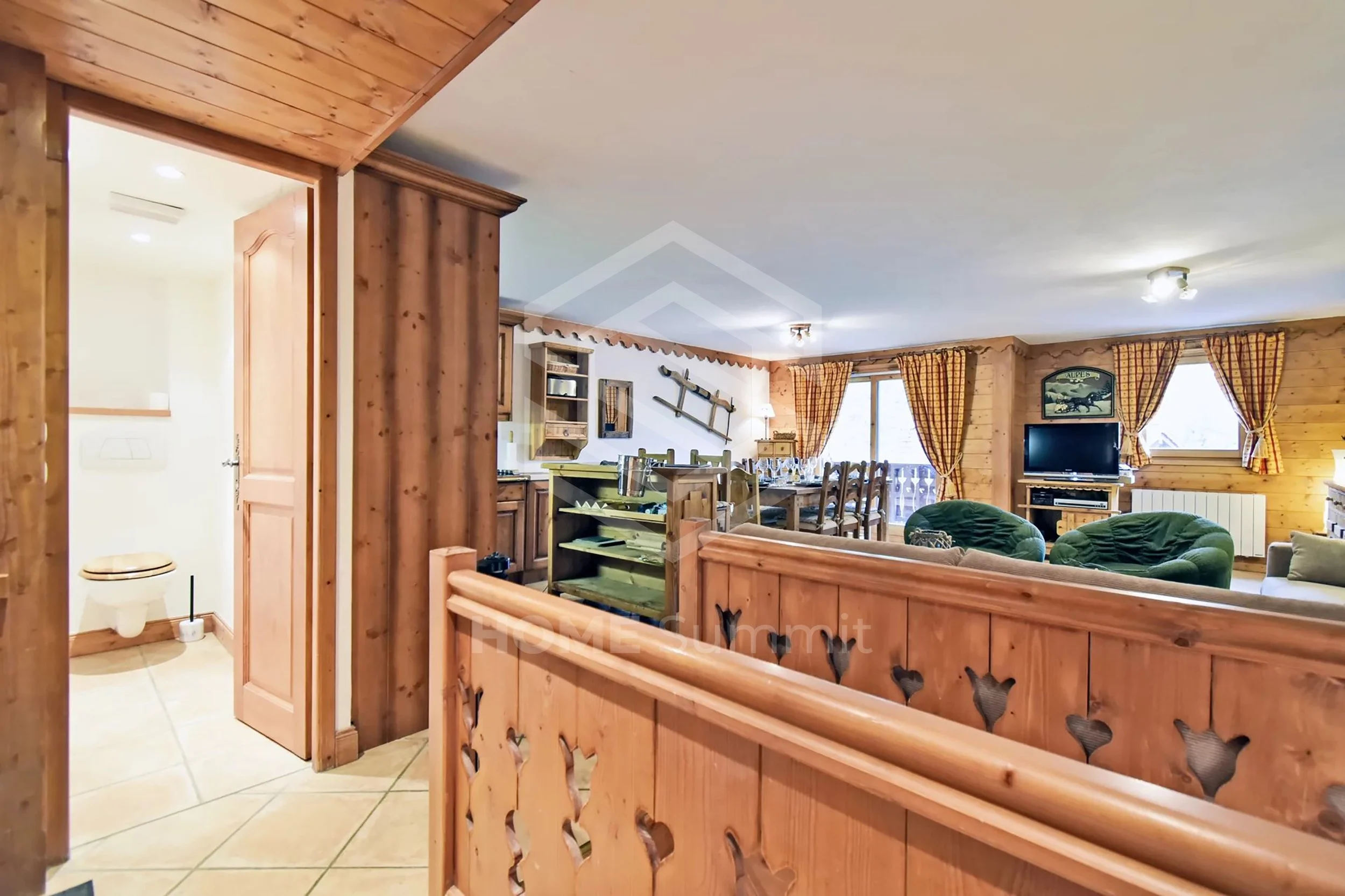 Salon avec canapés verts, télévision, salle à manger en bois, et murs en bois, vue depuis l'entrée, porte de salle de bain à gauche