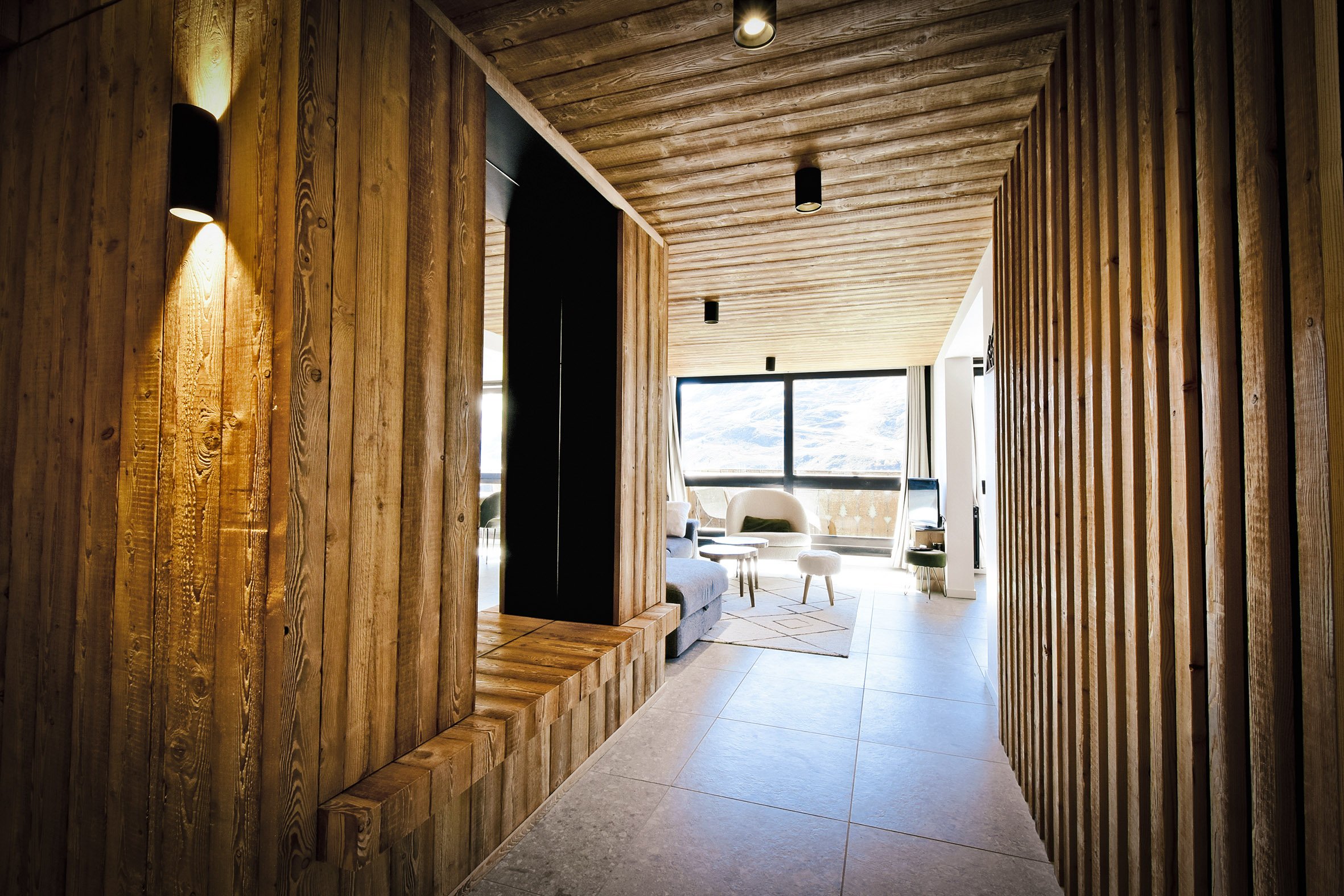 Salle de séjour moderne avec murs et plafond en bois, grande fenêtre laissant entrer la lumière naturelle, mobilier blanc et gris clair, tapis beige, télévision