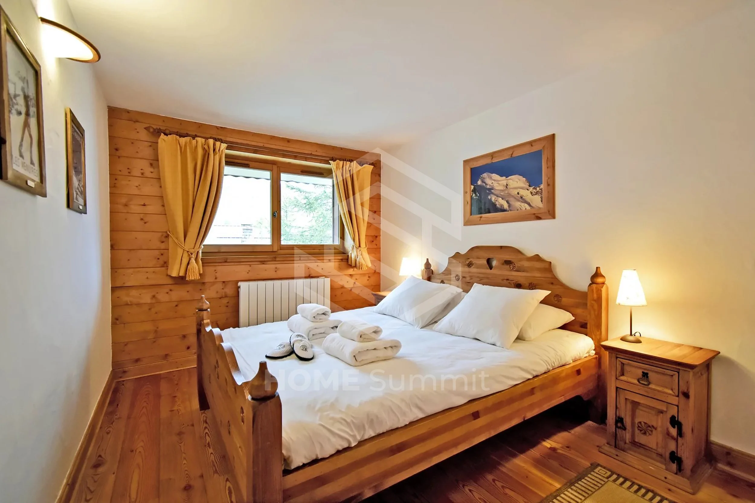 Chambre avec lit en bois, fenêtres avec rideaux jaunes, décor en bois, tableau de montagne au-dessus du lit, lampes de chevet, serviettes et chaussons sur le lit.