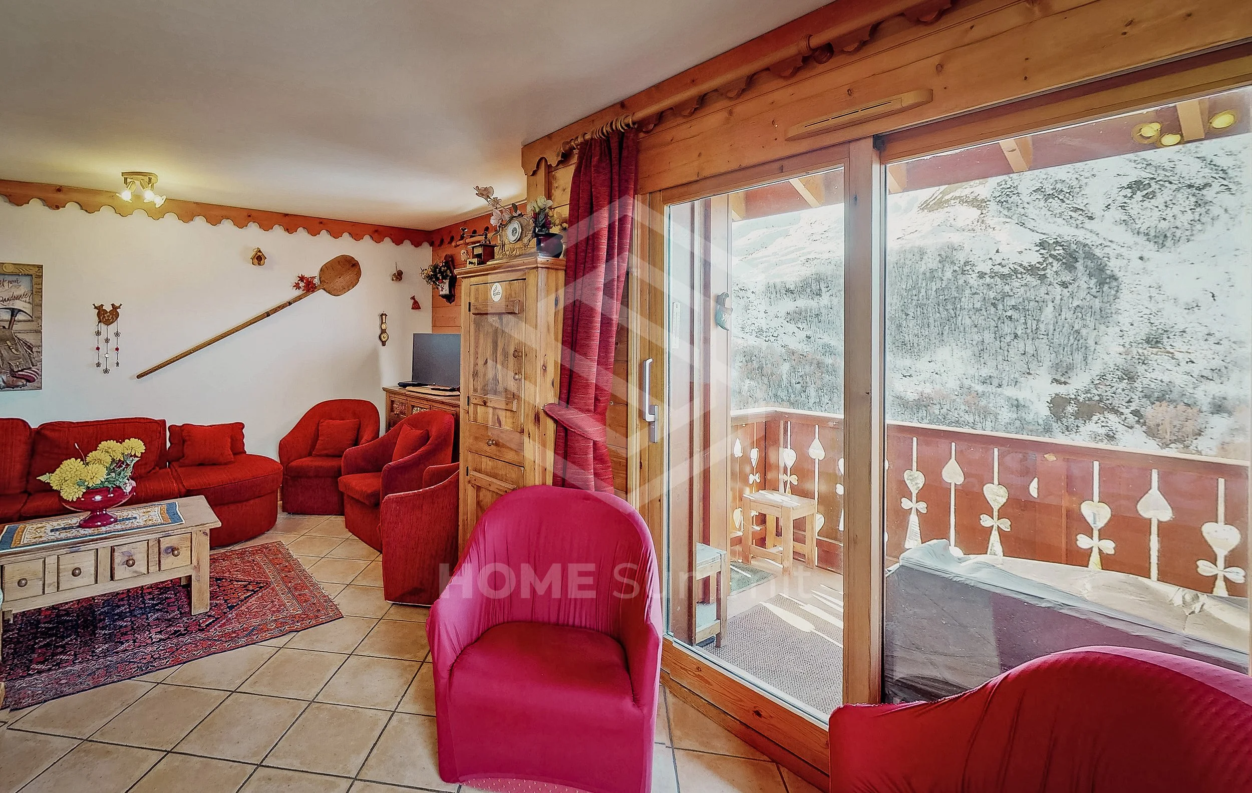 Salon chaleureux avec meubles rouges, y compris un canapé en forme de L, plusieurs fauteuils et une table en bois, décorations traditionnelles, mur en bois, grande fenêtre donnant sur une montagne enneigée.