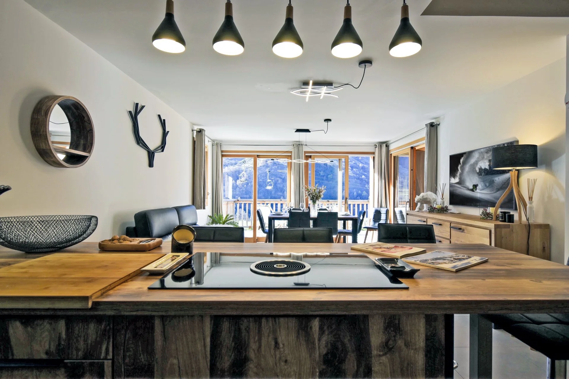 Intérieur d'une cuisine moderne avec un plan de travail en bois, un évier, un four à induction, et un coin repas en arrière-plan avec des fenêtres offrant une vue sur des montagnes.
