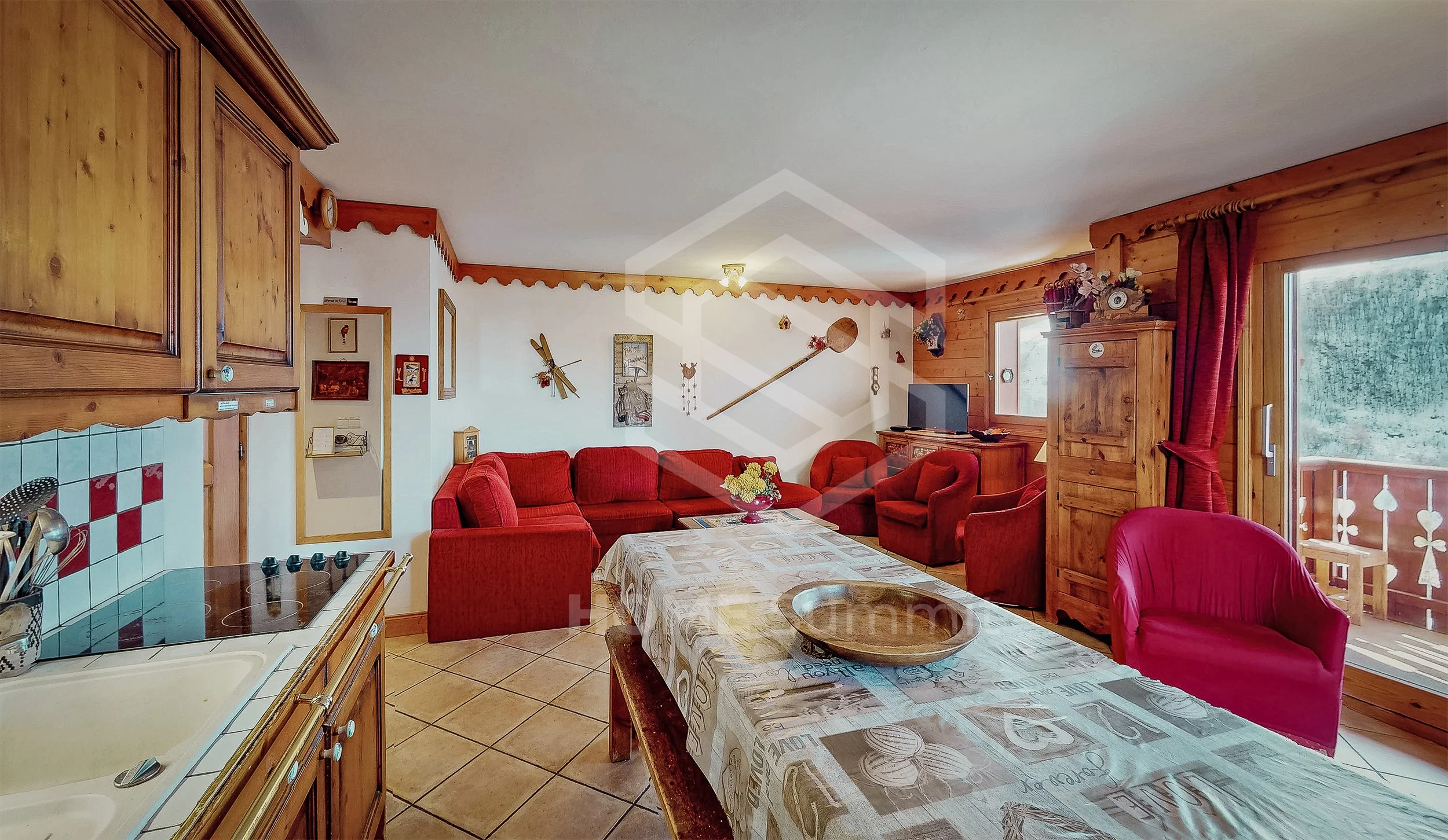 Salon et cuisine dans une maison en bois, meubles en bois, canapé rouge, fauteuils rouges, table avec nappe décorée, télévision, fenêtres avec volets en bois, décorations murales, balcon.