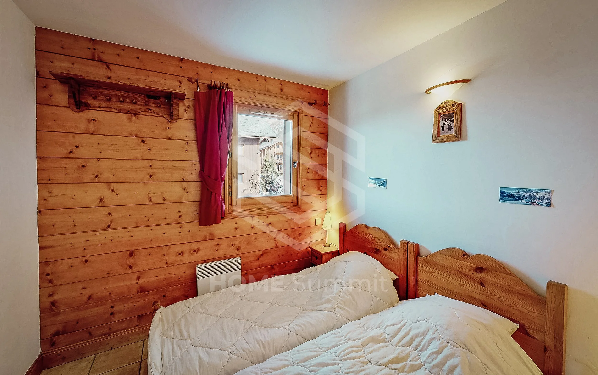 Chambre avec deux lits en bois, une fenêtre avec rideau rouge, mur en bois et mur blanc avec des photos, petit lampe de chevet, sol en carrelage.