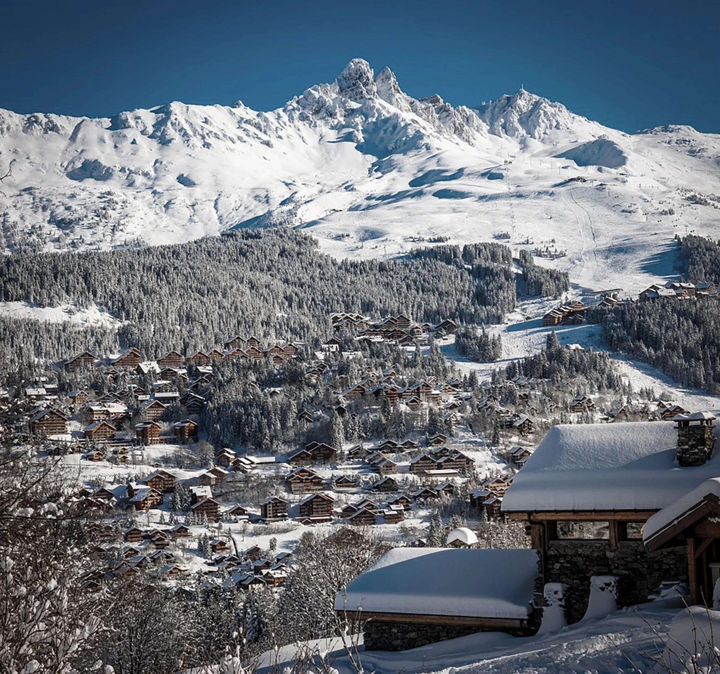 Vue d'une station de montagne enneigée avec de nombreux chalets en bois, entourée de forêts et de hautes montagnes enneigées sous un ciel clair.