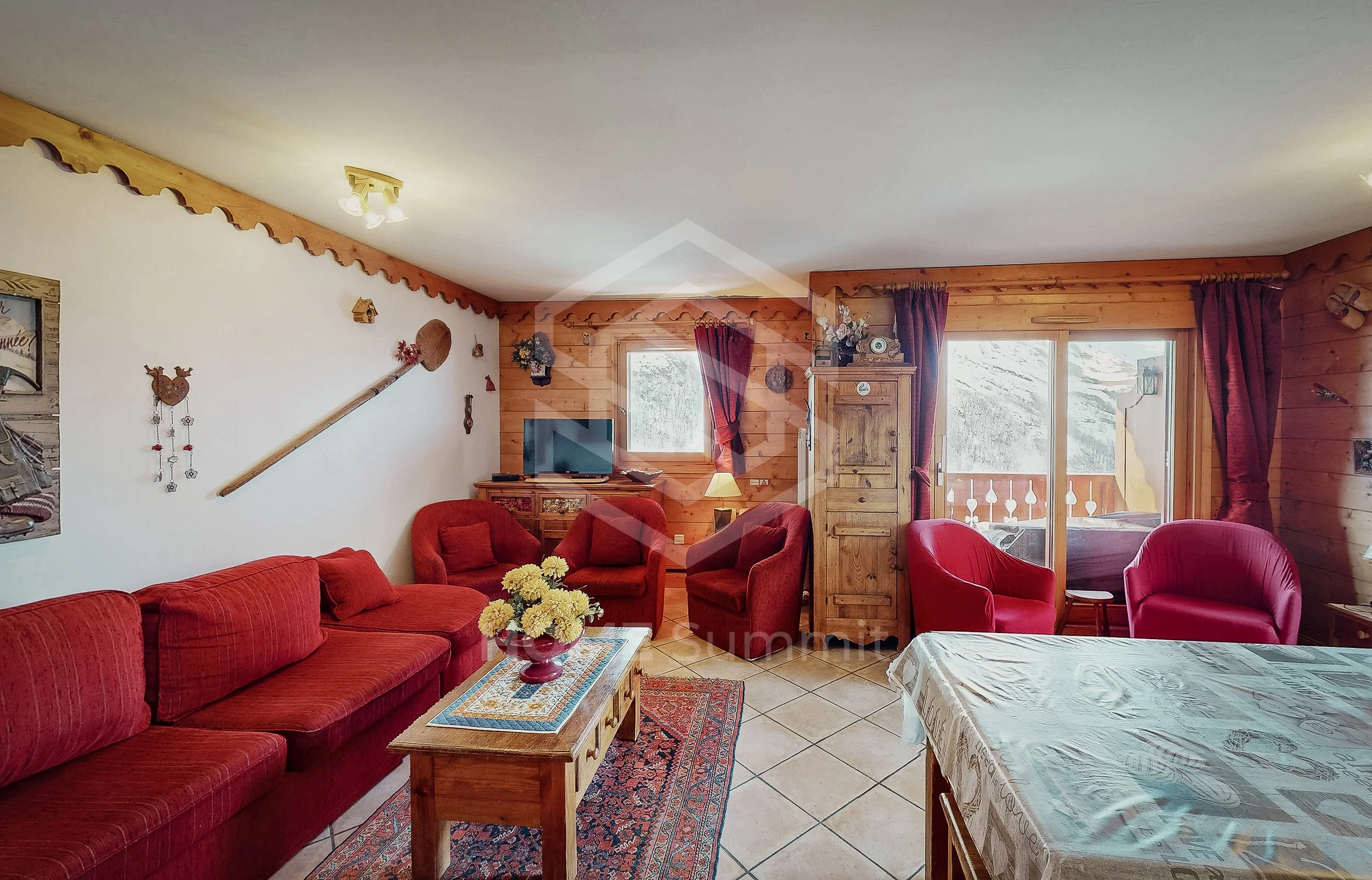 Salon de chalet avec canapé rouge, fauteuils rouges, télévision, cheminée, meubles en bois, balcon avec vue sur la montagne