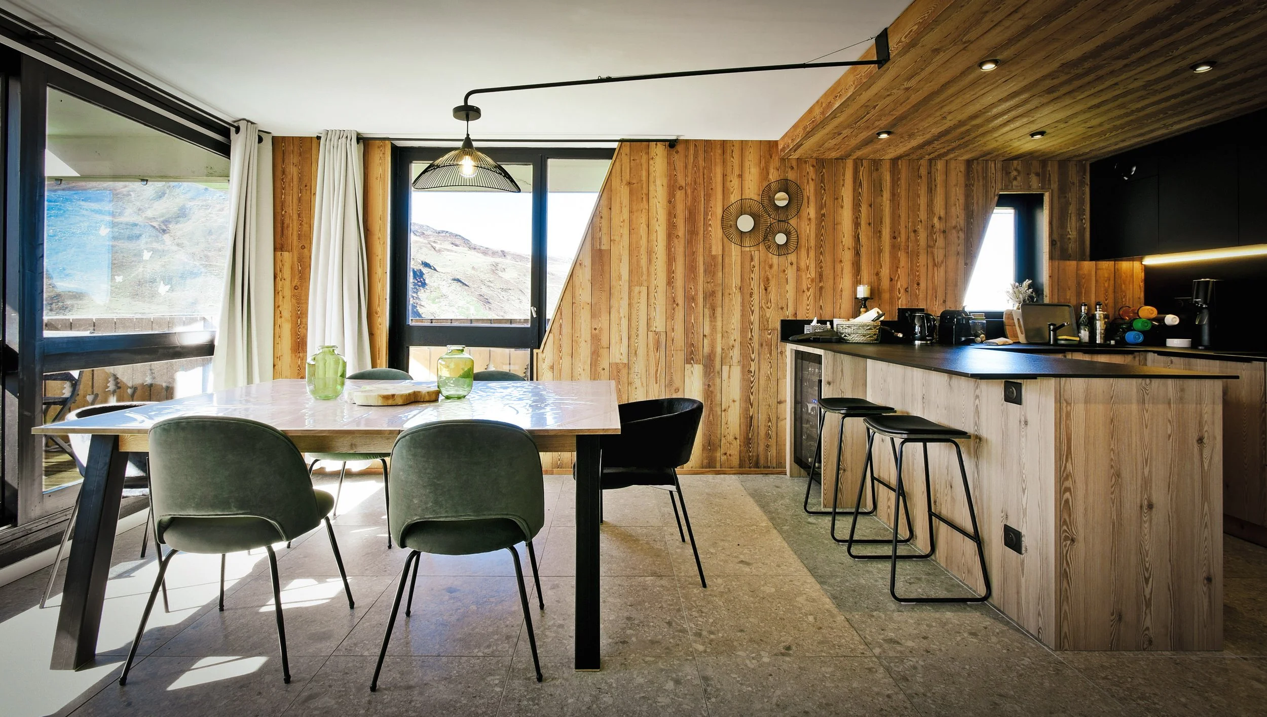 Intérieur d'une cuisine moderne avec un mur en bois clair, un plan de travail en bois clair, un coin repas avec une table en bois et plusieurs chaises vertes et noires, grandes fenêtres laissant entrer la lumière naturelle, décorations murales rondes, et des ustensiles de cuisine