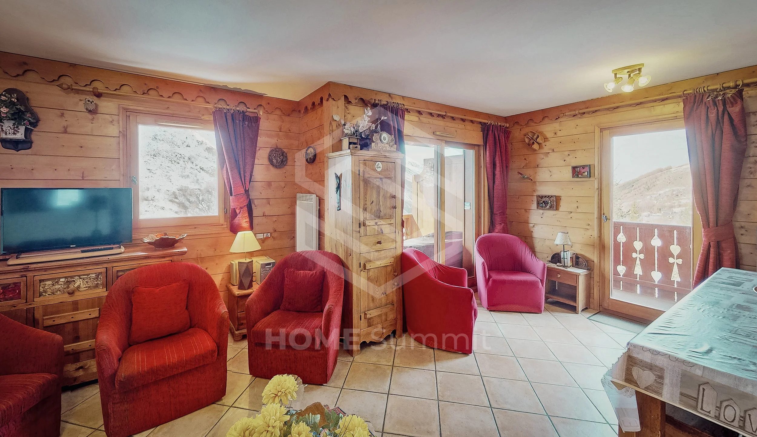 Salon de chalet en bois avec mobilier rouge, télévision, fenêtres avec vue sur montagne, porte-fenêtre avec balcon, sol en carreaux, décoration chaleureuse.
