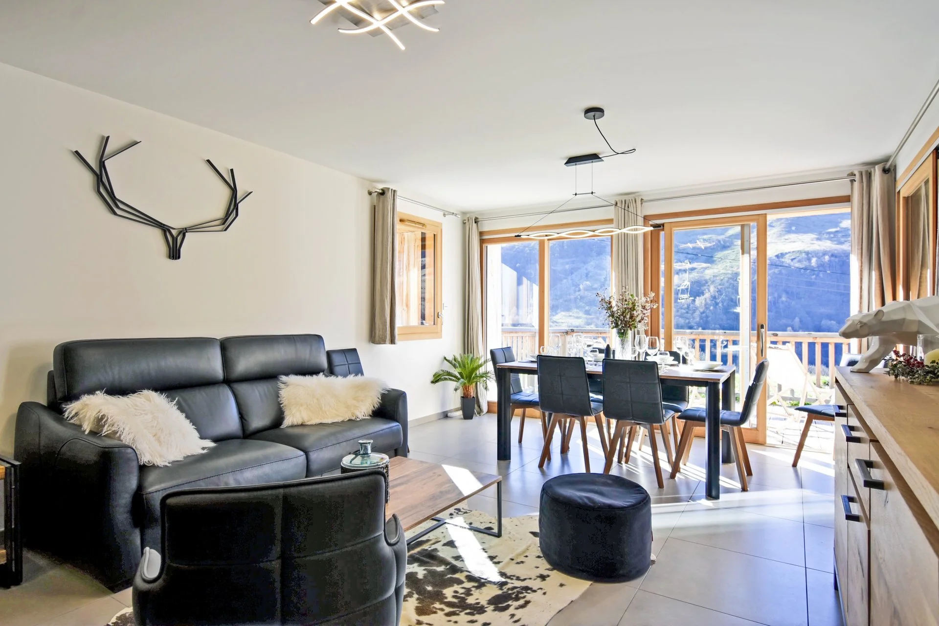 Salon avec canapé en cuir noir, table basse, fauteuils, table à manger avec chaises, grande fenetre offrant vue sur montagnes, décoration moderne