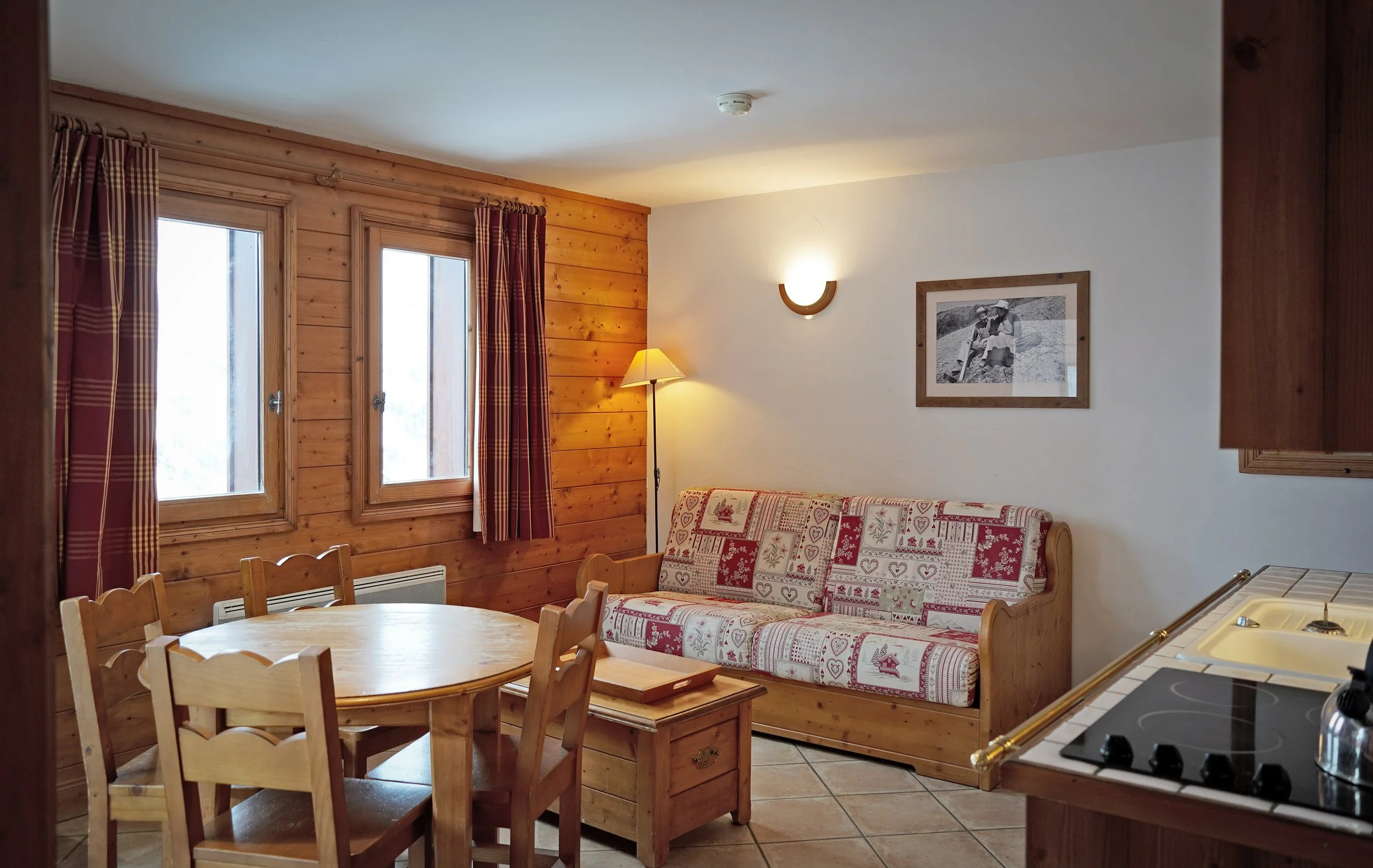 Intérieur d'une pièce de style chalet en bois, avec fenêtres, une table en bois, des chaises, un canapé avec housse à motifs de cœurs et maisons, une lampe sur pied, un tableau noir et blanc, et un mur blanc avec un éclairage mural
