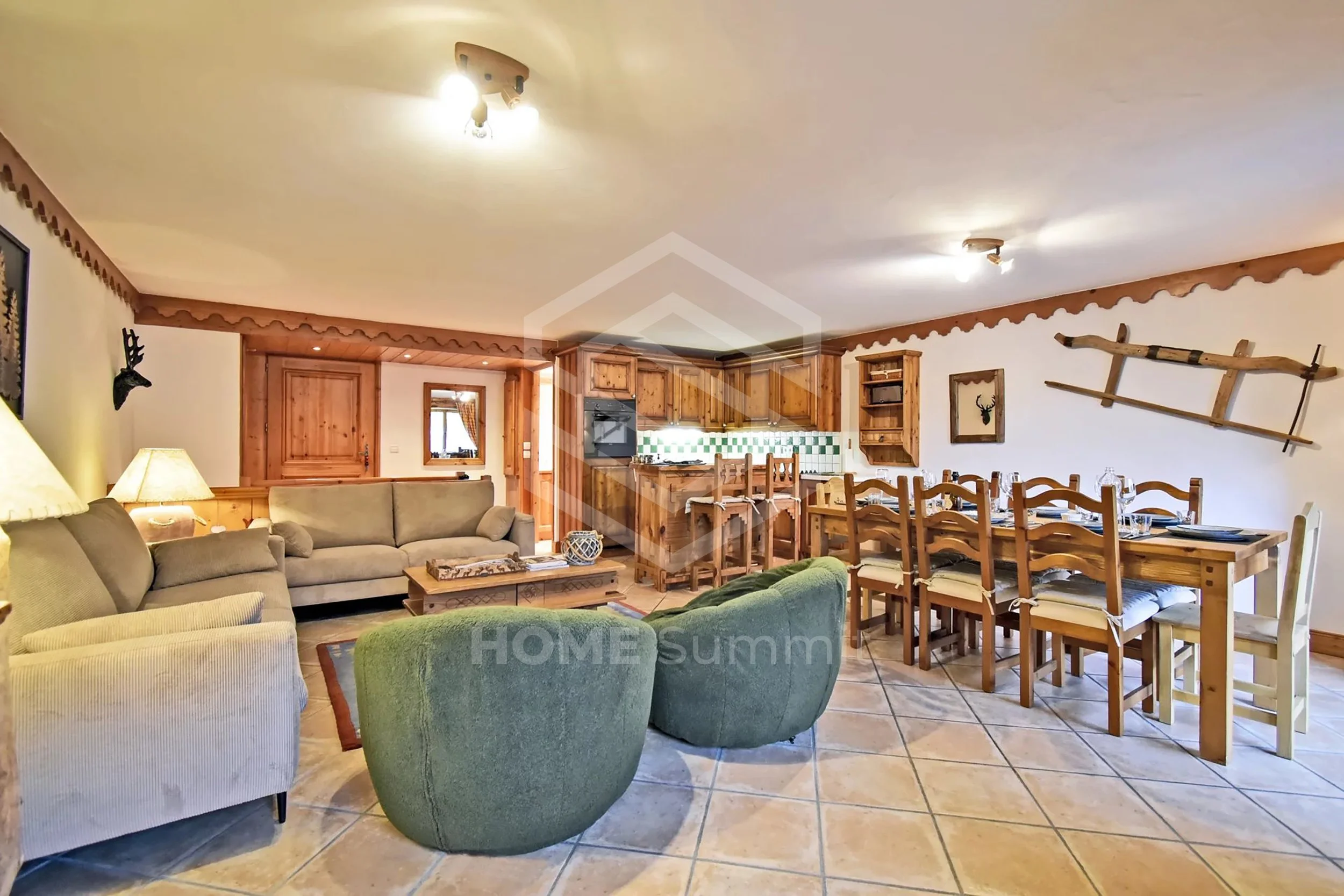 Salon et coin repas dans un chalet en bois, avec canapé beige, fauteuil vert, grande table en bois avec chaises, cuisine en arrière-plan, décor rustique