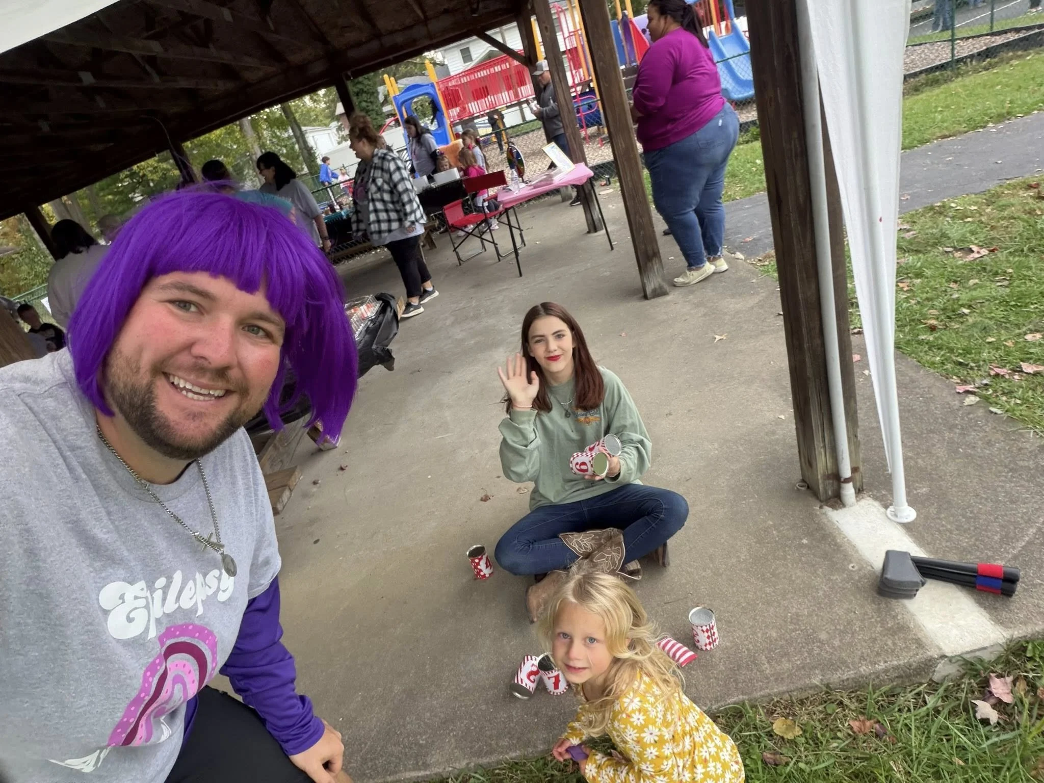 A group of people at an outdoor gathering, with a man wearing a purple wig in the foreground, a woman sitting cross-legged on the ground waving, and a young girl in a yellow dress with white flowers. There are tables, tents, and a playground in the b