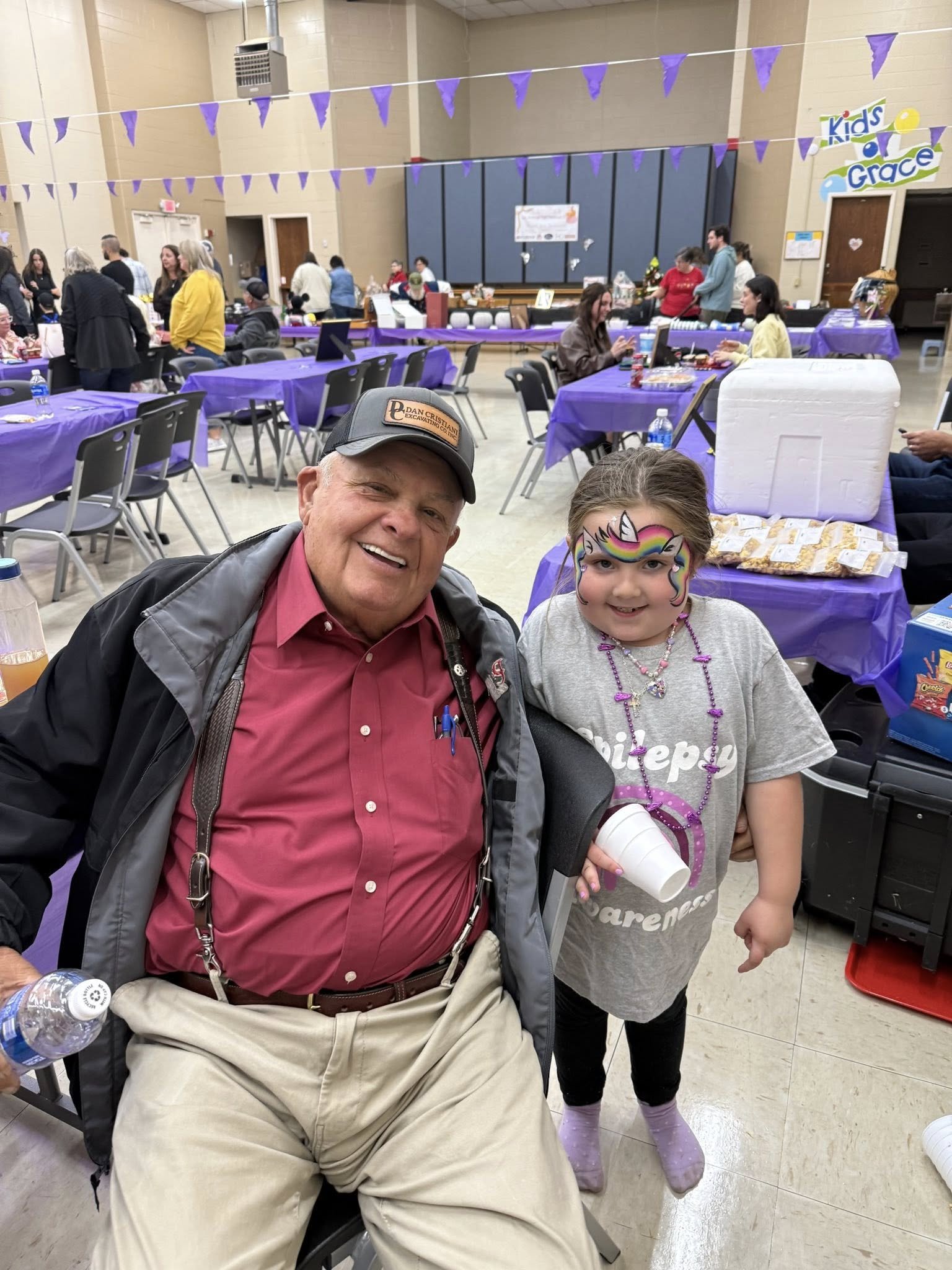 Smiling elderly man and young girl in unicorn face paint at a community event with purple tablecloths, balloons, and other attendees.