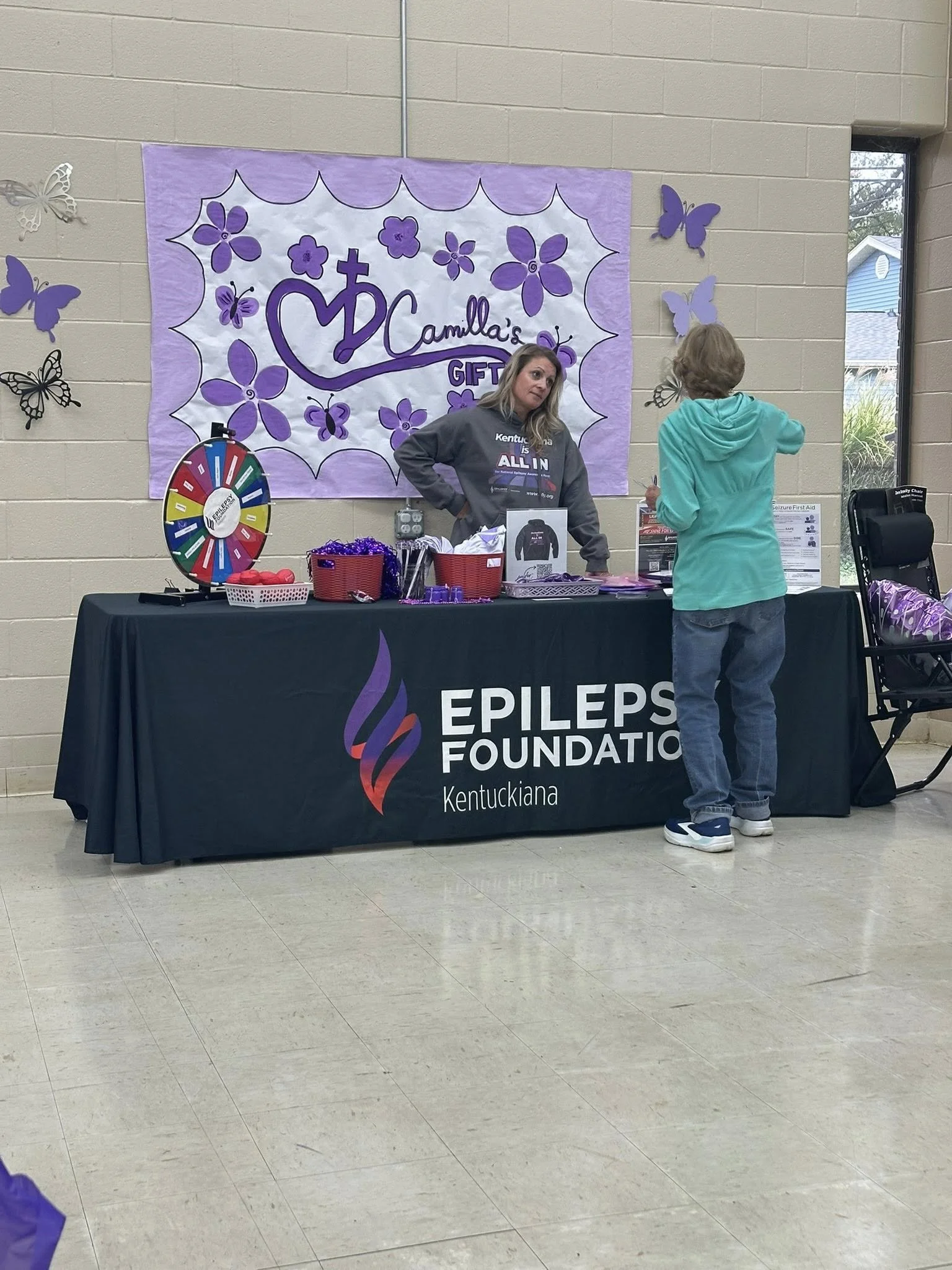 A woman at an epilepsy awareness event booth for the Epileps Foundation Kentucky, with a man standing in front of her. The booth has a black tablecloth with the foundation's logo, a prize wheel, purple decorations, and informational materials.