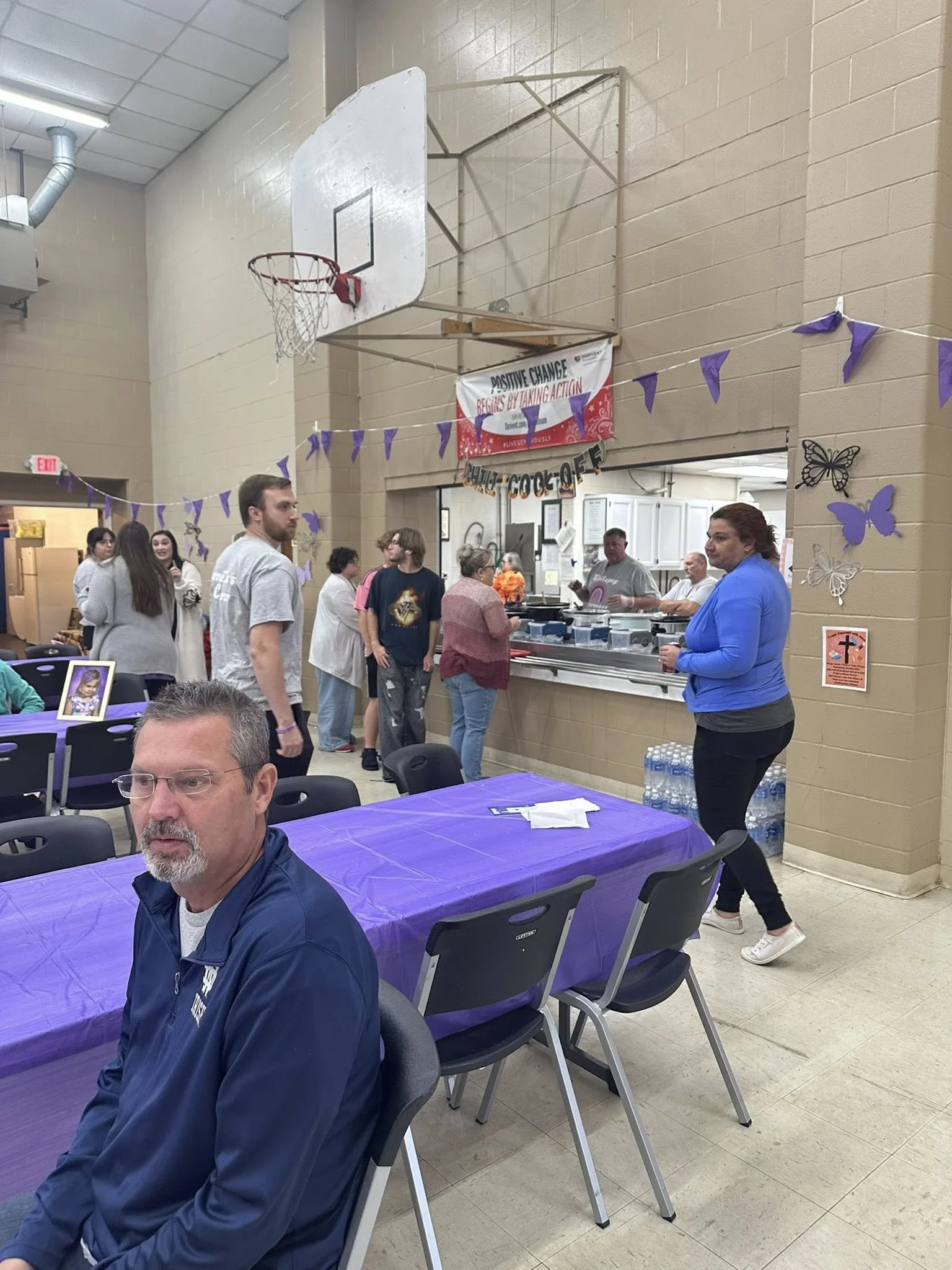 People gathered in a community center with a serving line, purple decorations, and tables covered with purple tablecloths, participating in a social event.