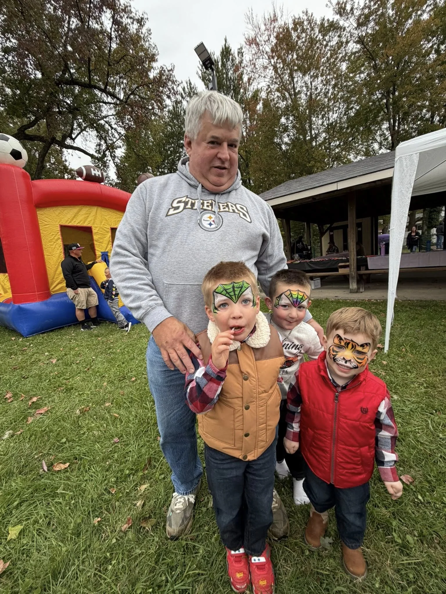 A man in a gray Steelers hoodie with three children with face paint, two of them with spider web designs and one with a tiger face, at an outdoor event on a grassy area with trees and a bounce house in the background.