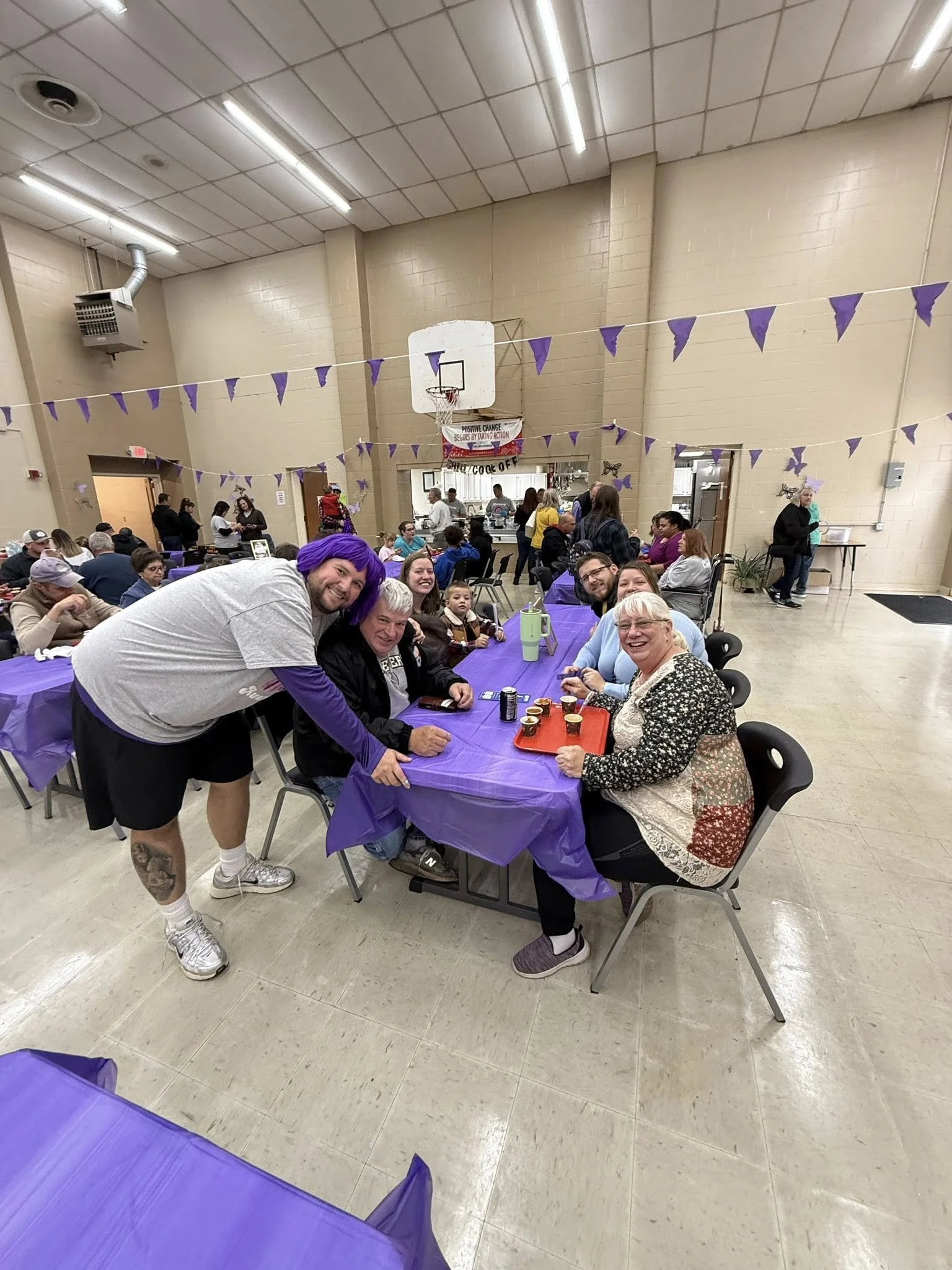 Group of six people sitting and standing around a purple table at a community gathering or celebration in a large hall with purple decorations.