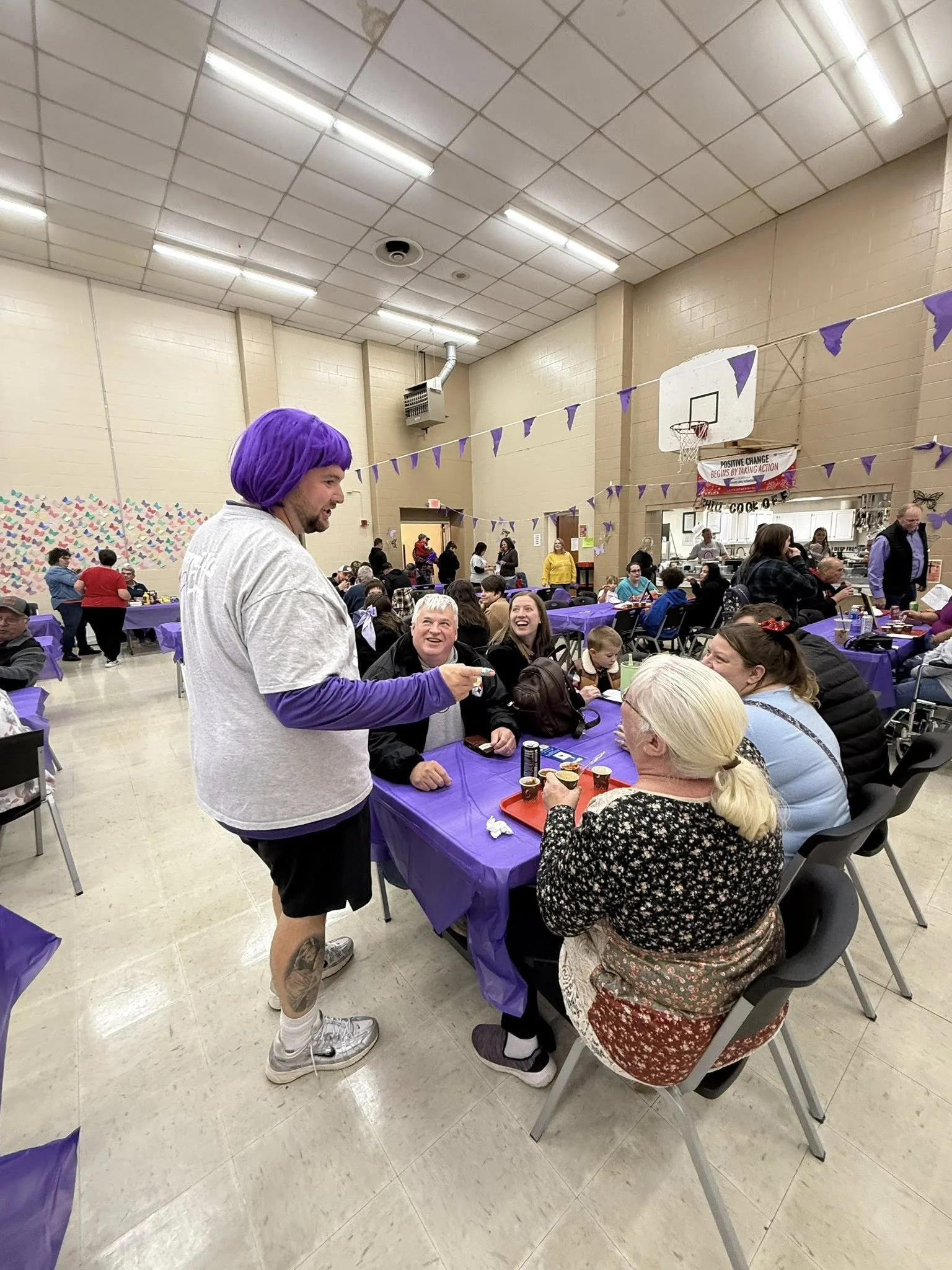 A man with purple hair and purple sleeves serving food to a group of smiling people at a community event in a gymnasium decorated with purple banners.