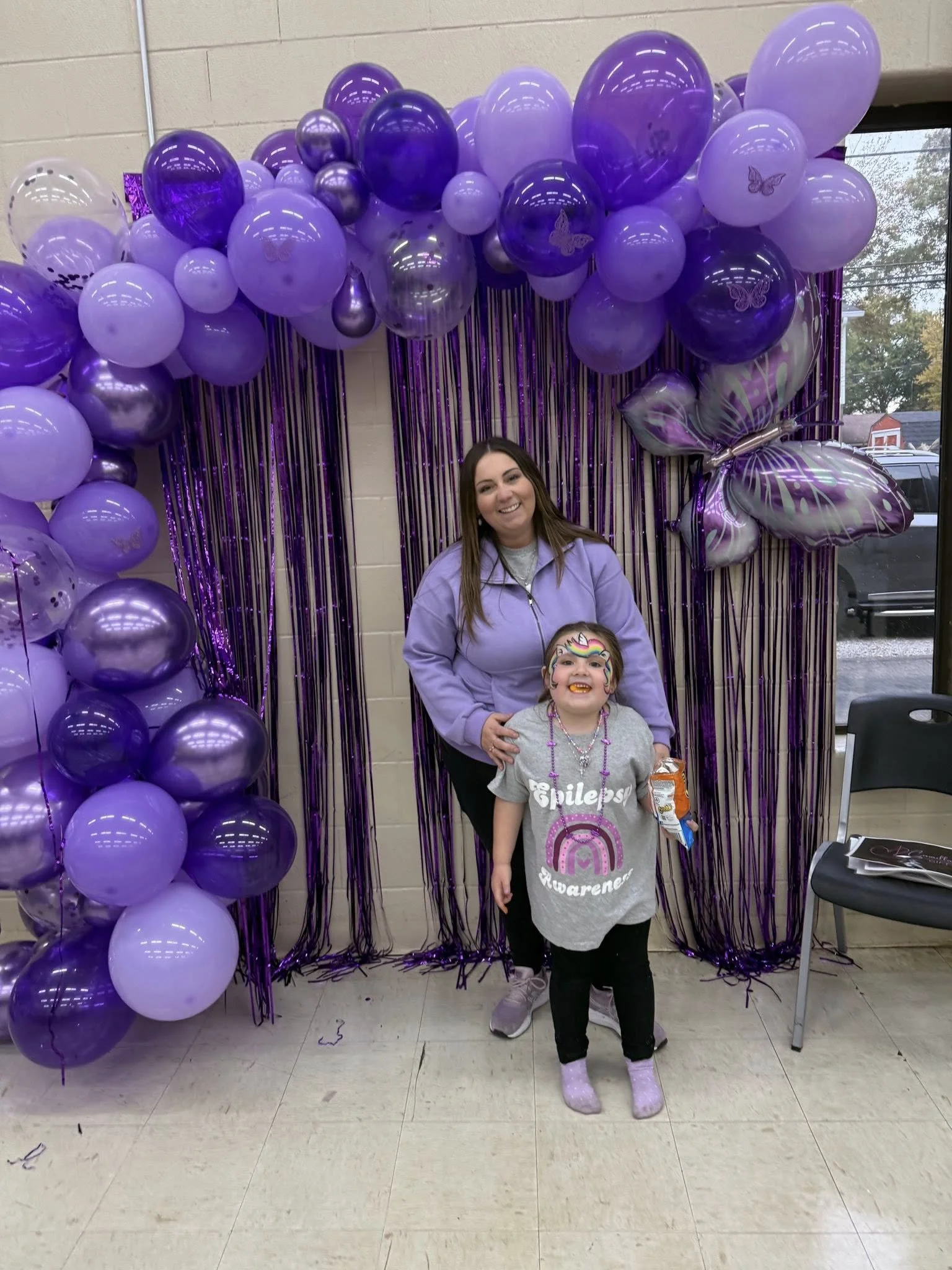A woman and a young girl standing in front of purple balloon decorations at a celebration. The girl has face paint, is smiling, and holding a snack.