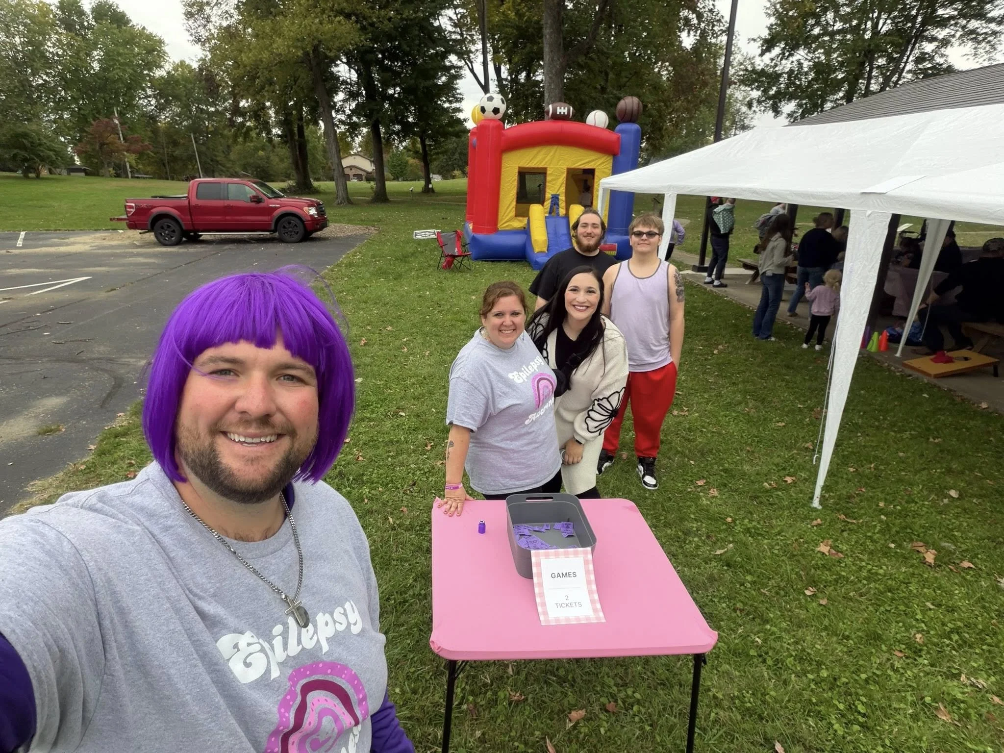 Group of people at outdoor event, a man wearing a purple wig and gray T-shirt taking selfie, others standing behind a pink table with tickets, with an inflatable bounce house and children in the background.