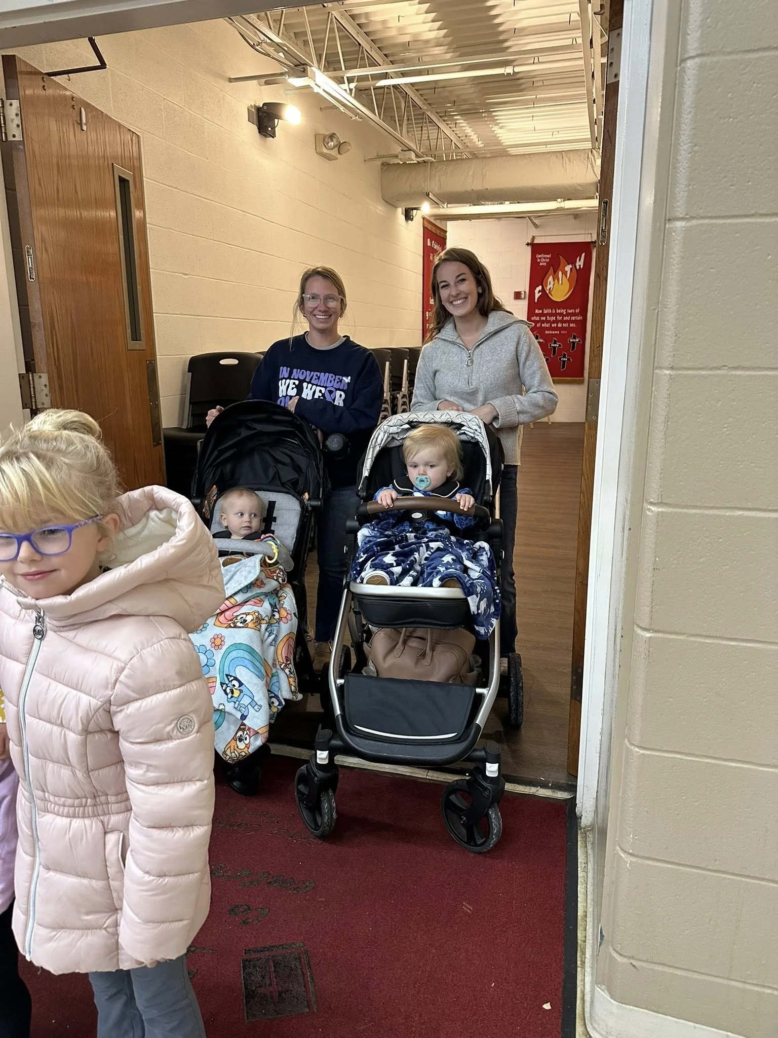 Two women and three young children, two in strollers, standing in a hallway at a church or community center.