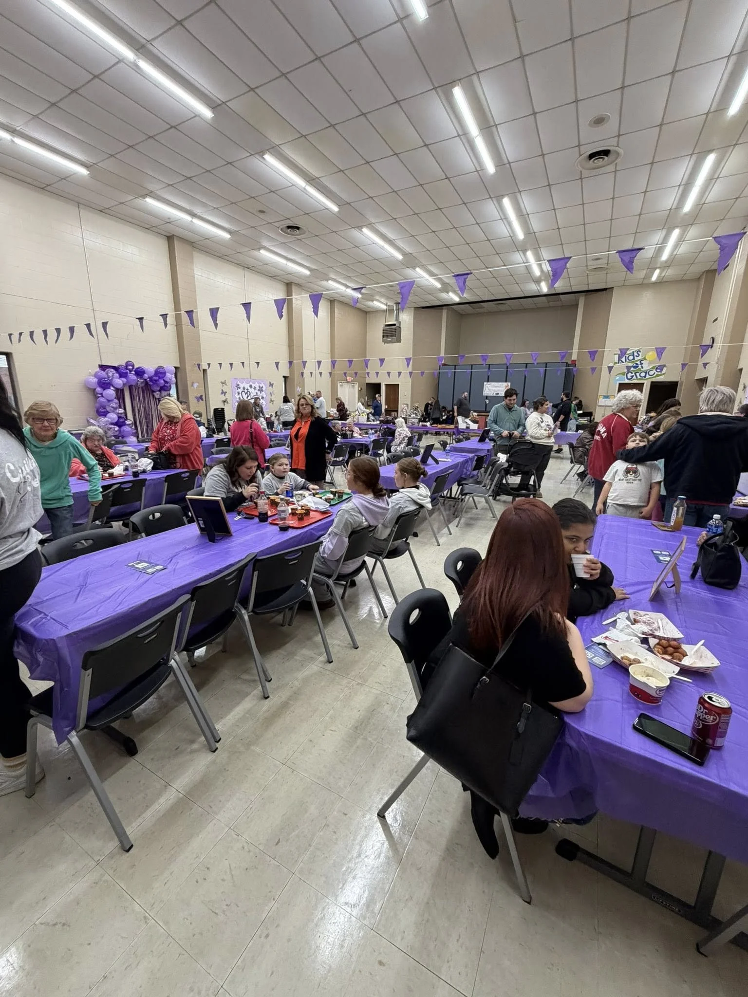 Community gathering in a hall decorated with purple banners and balloons, with tablecloths and tables set up for food and socializing.
