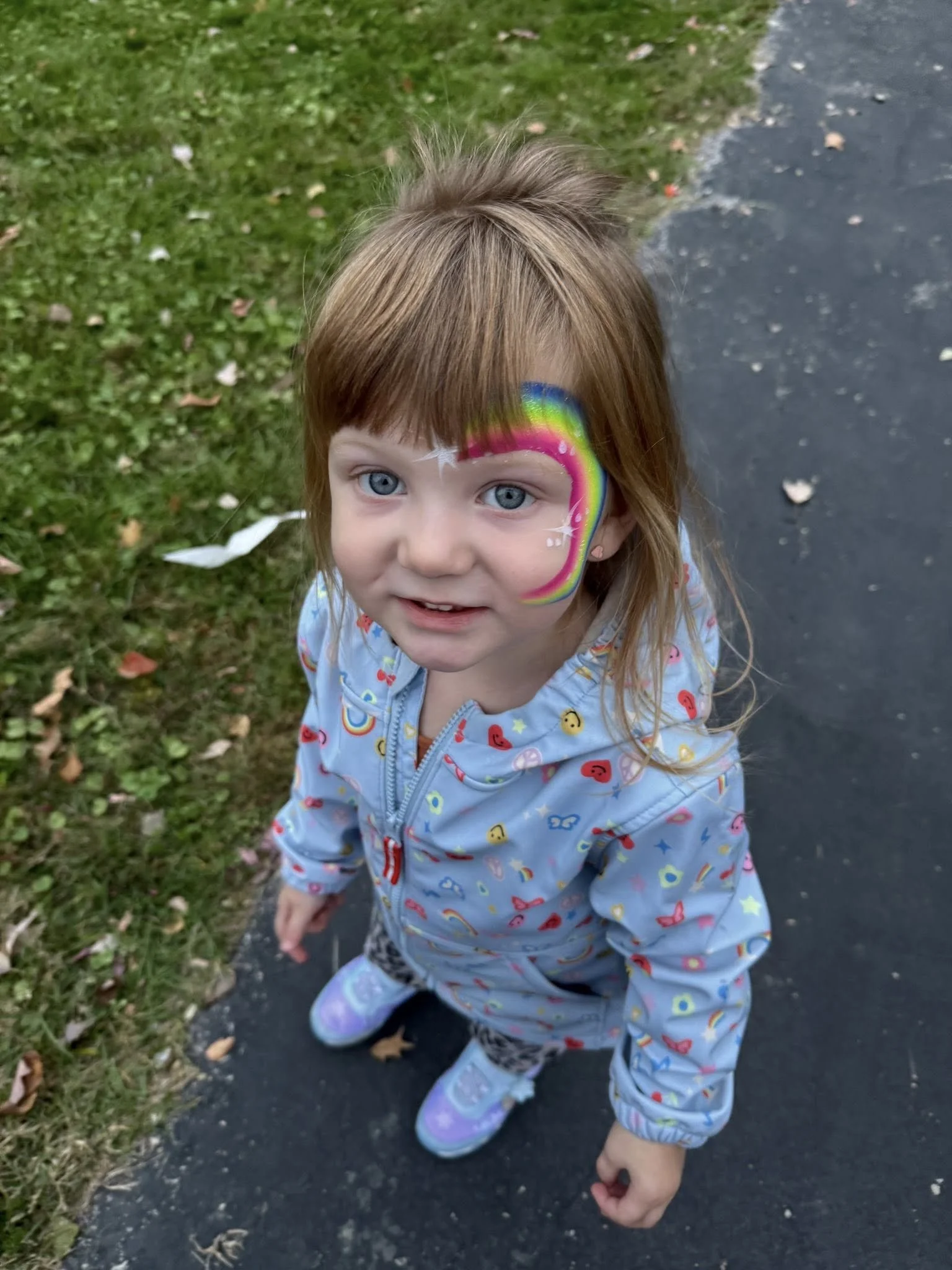 A young girl with blue eyes and shoulder-length light brown hair standing on a sidewalk, with rainbow face paint on her face.