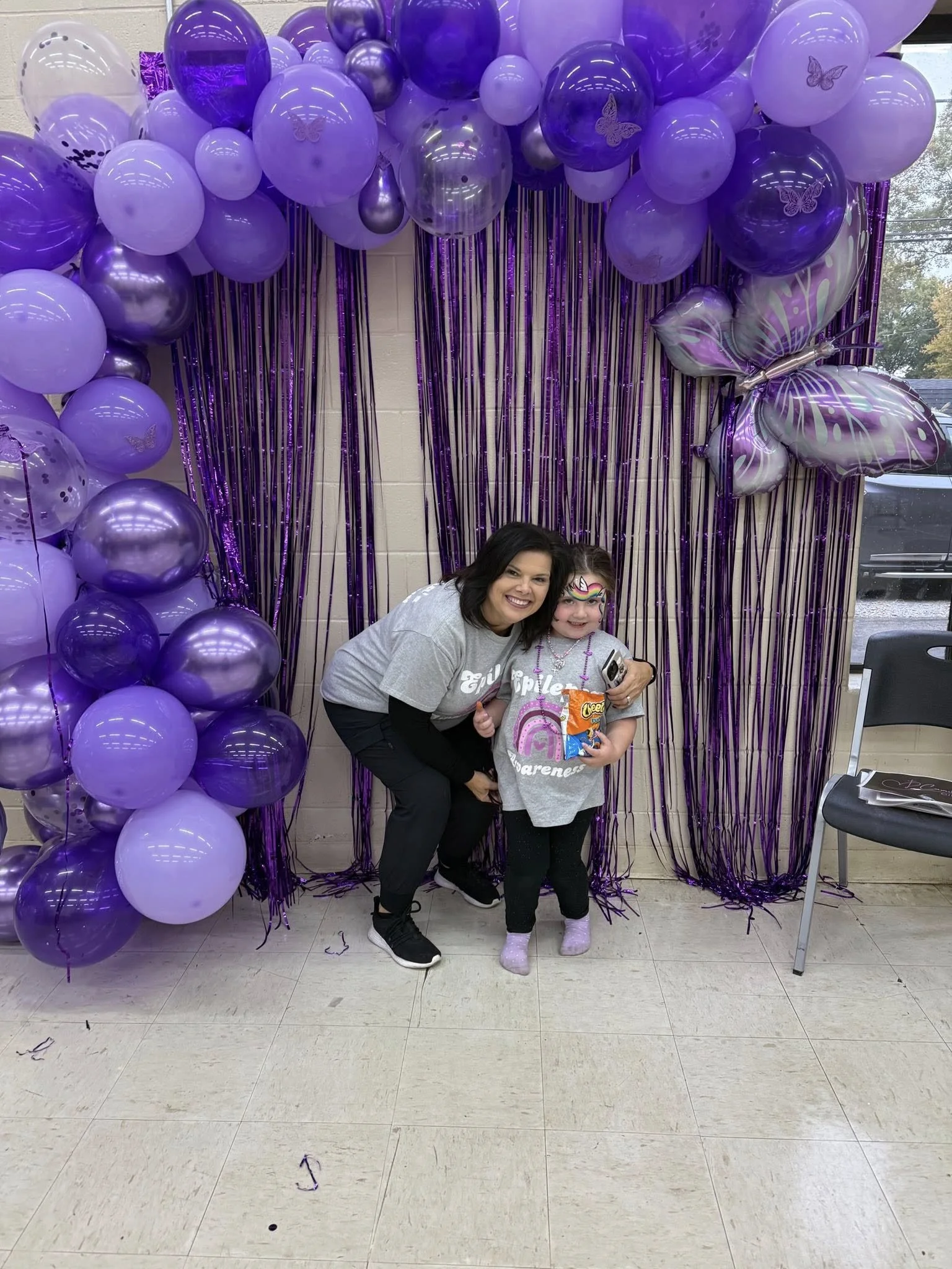 A woman and a girl dressed in gray shirts with rainbow design posing and smiling in front of purple balloons and metallic butterfly balloon decorations at a celebration event.