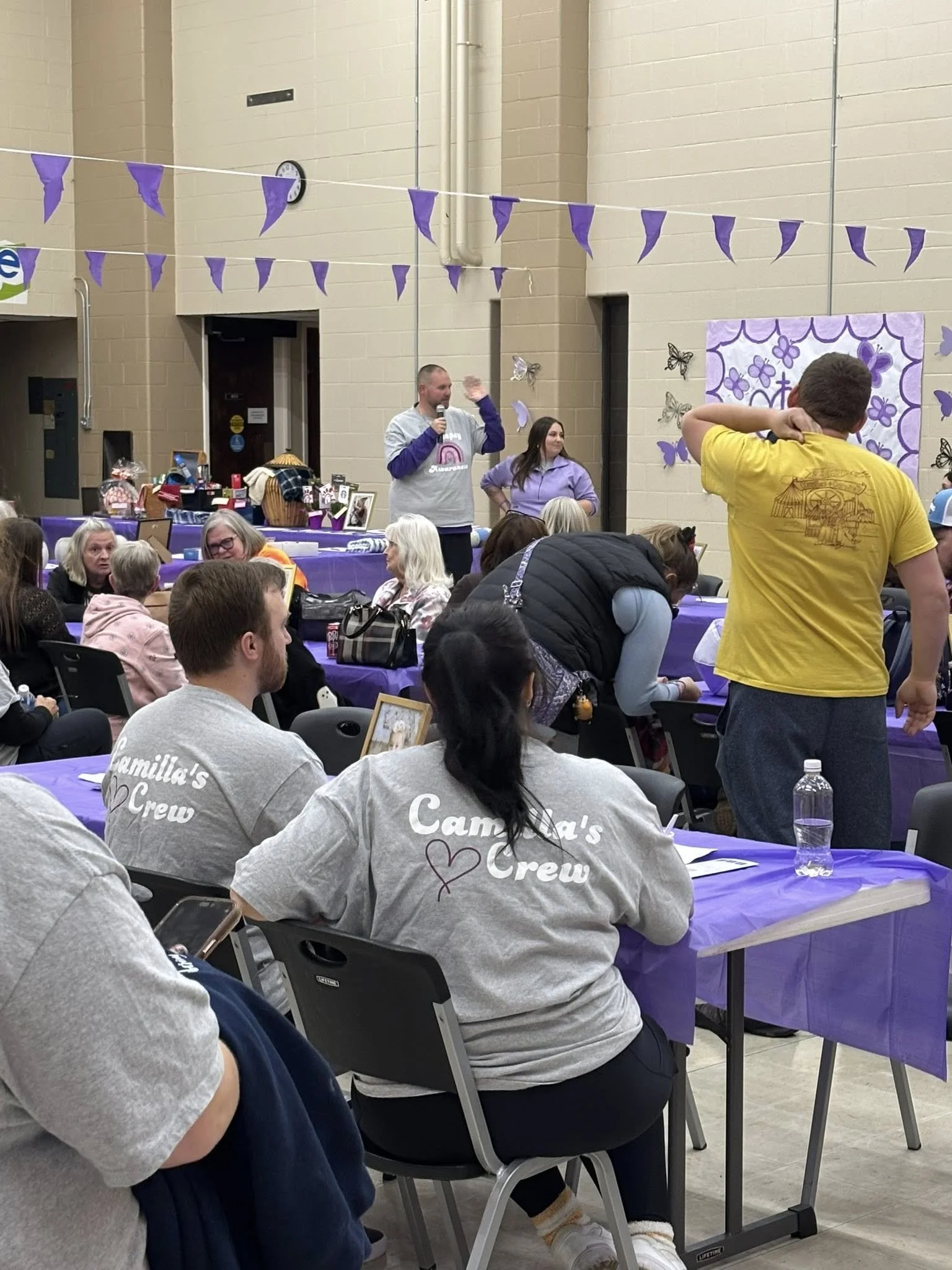 People attending an indoor event with purple decorations, some wearing matching gray shirts that say 'Camilia's Crew,' seated at tables with purple tablecloths, while a man with a microphone stands addressing the crowd.
