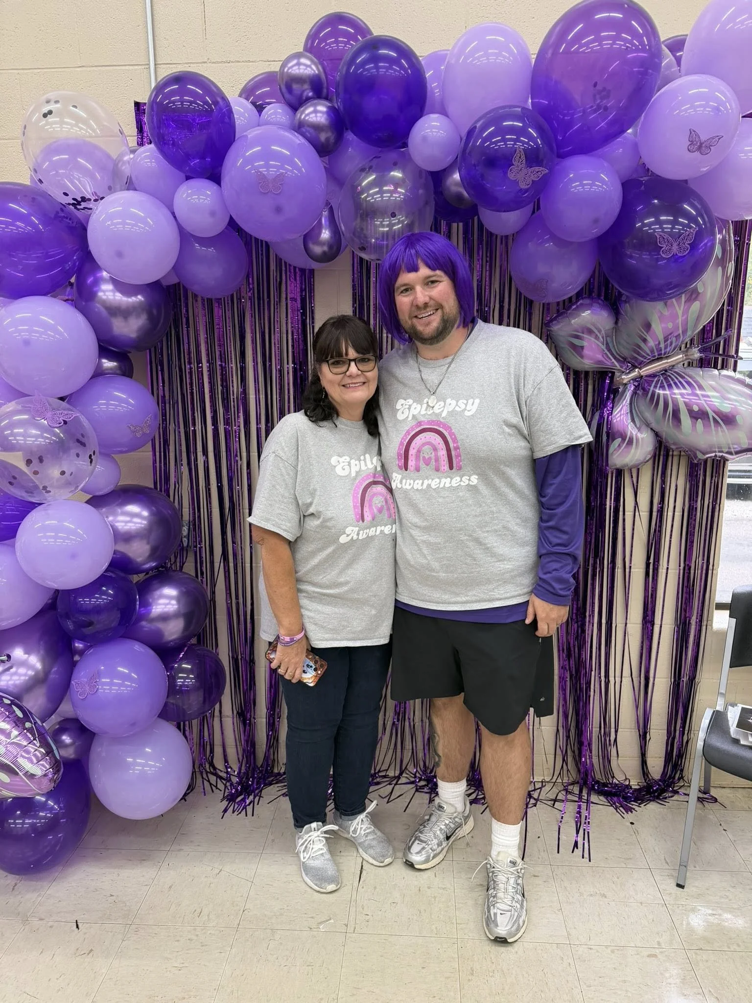 Two people standing in front of a purple balloon arch with metallic and butterfly-patterned balloons, wearing gray T-shirts with 'Epilepsy Awareness' and rainbow logos, smiling.