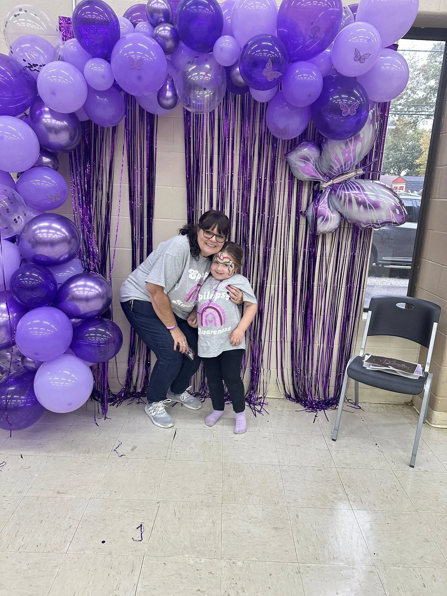 A woman and a young girl with face paint, smiling and posing in front of purple balloons, metallic streamers, and a large butterfly-shaped balloon, at a celebration or party.