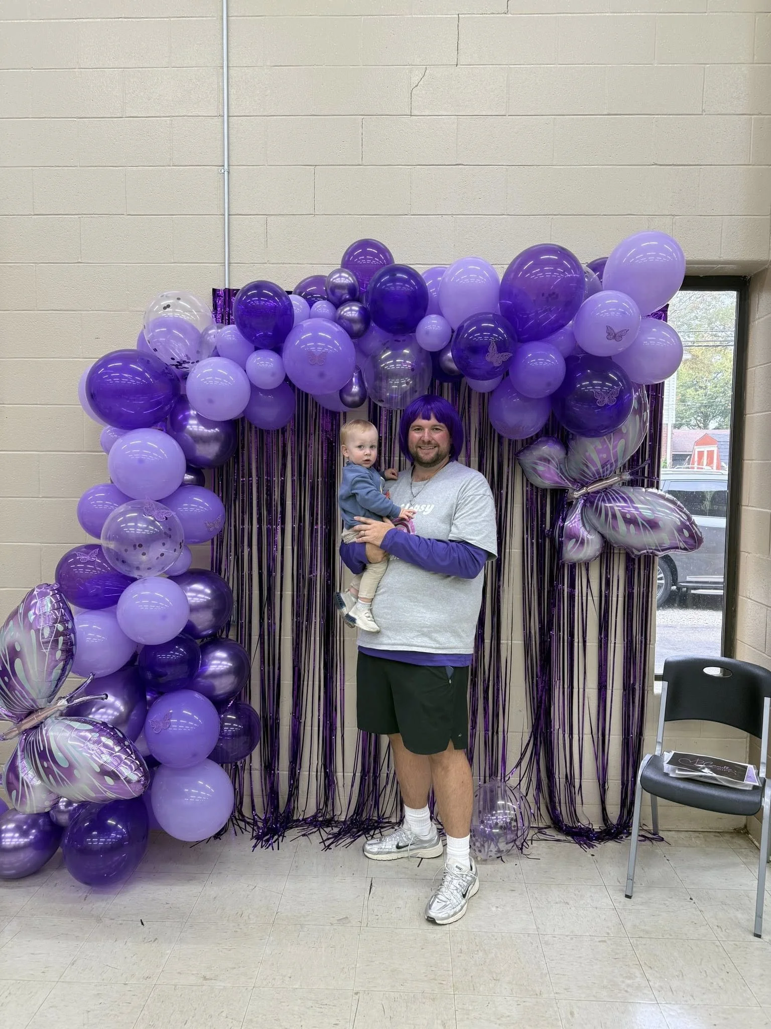 A man with a purple wig holding a child is standing in front of a purple balloon arch with butterfly and butterfly-themed balloons, purple streamers, and a window showing an outdoor scene.