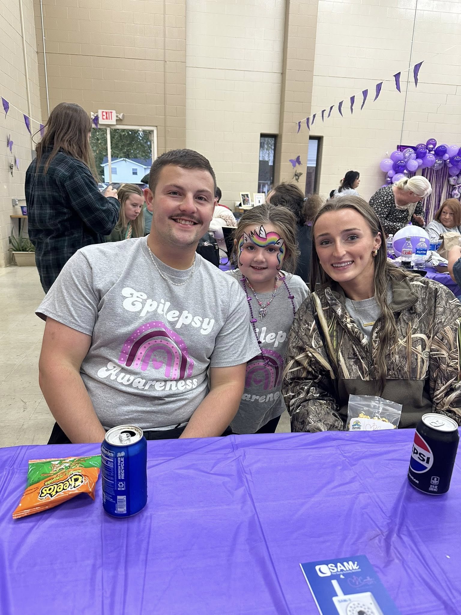 Three people sitting at a table decorated for epilepsy awareness, with purple decorations in the background. The man and woman are smiling, and the girl in the middle has face paint of a unicorn. They are wearing matching gray shirts that say 'Epilep
