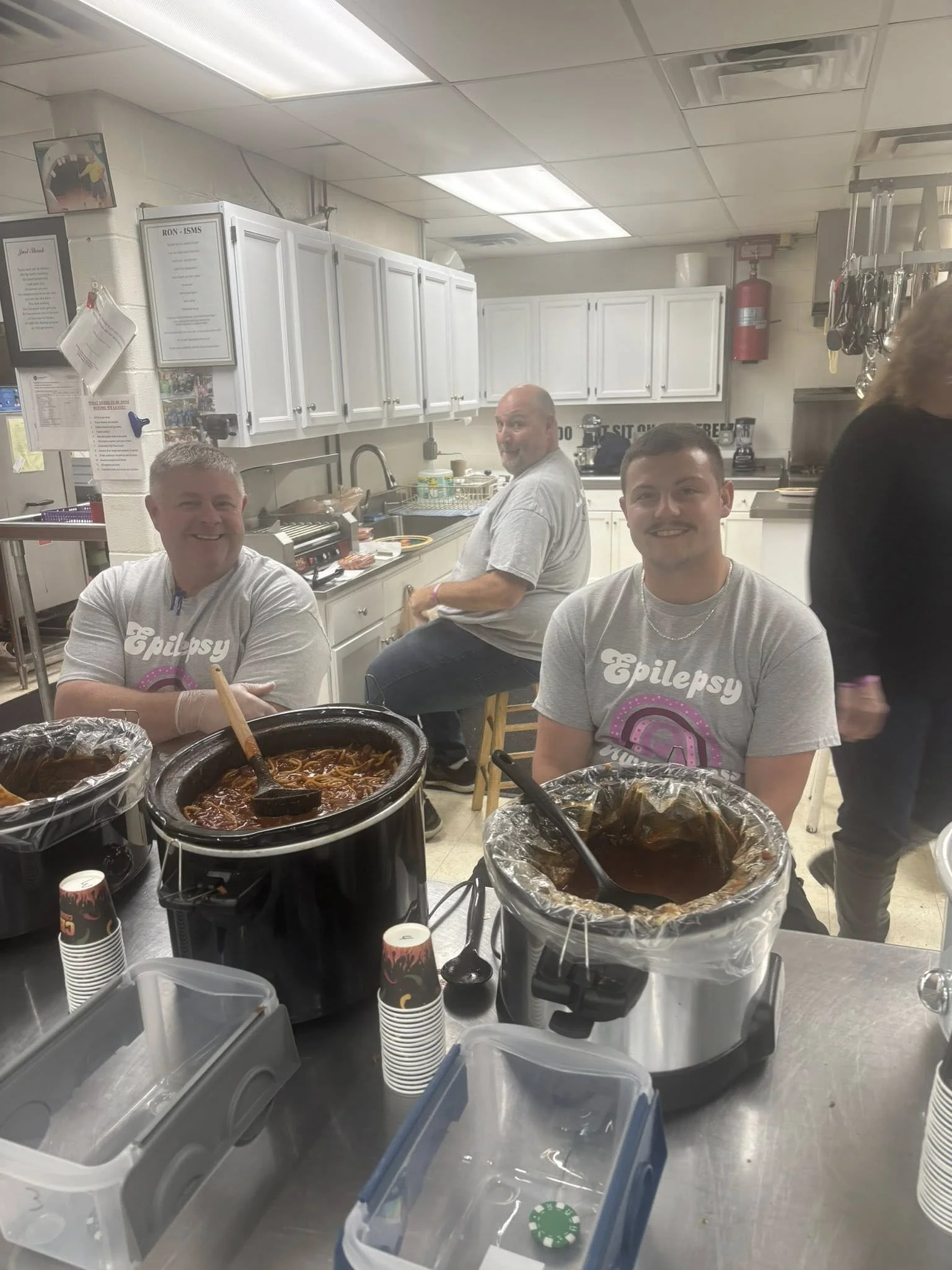 Three men in gray T-shirts with 'Epilepsy' logo sitting at a table with food and drinks, one man in the background, kitchen setting.