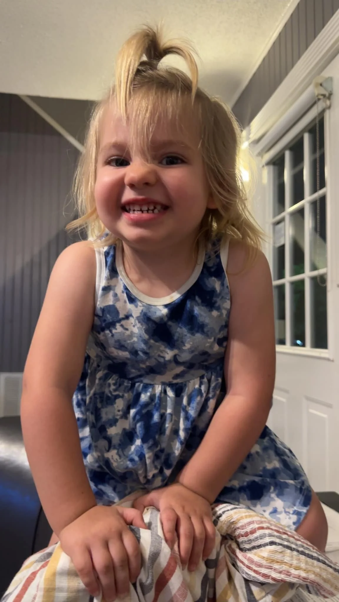A young girl with blonde hair, smiling and showing her teeth, wearing a blue and white patterned dress, leaning over a striped cloth, indoors with a window in the background.