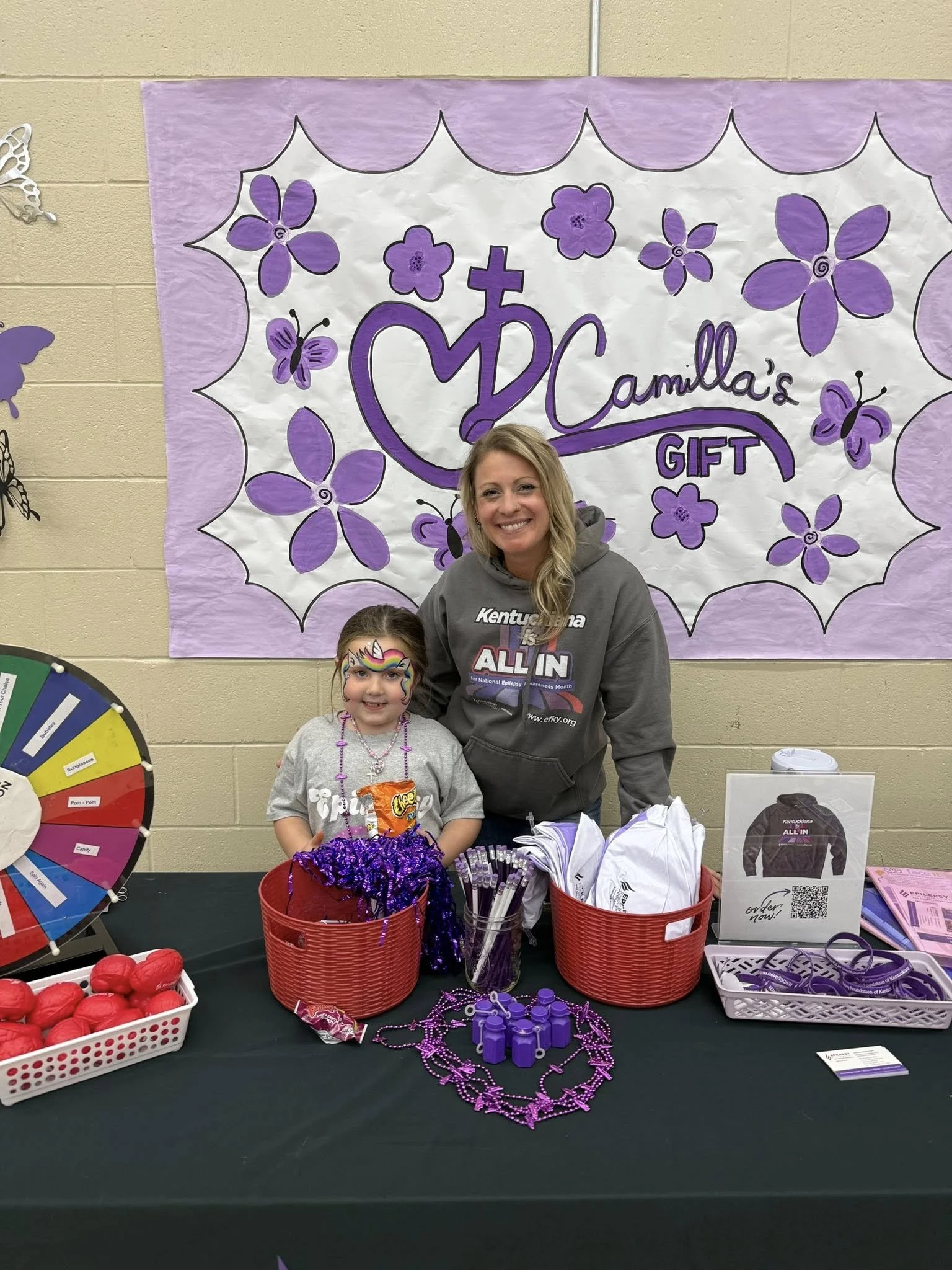 A woman and young girl standing behind a table with purple-themed items at Camilla's gift booth, with a purple and white floral sign reading "Camilla's Gift" behind them.