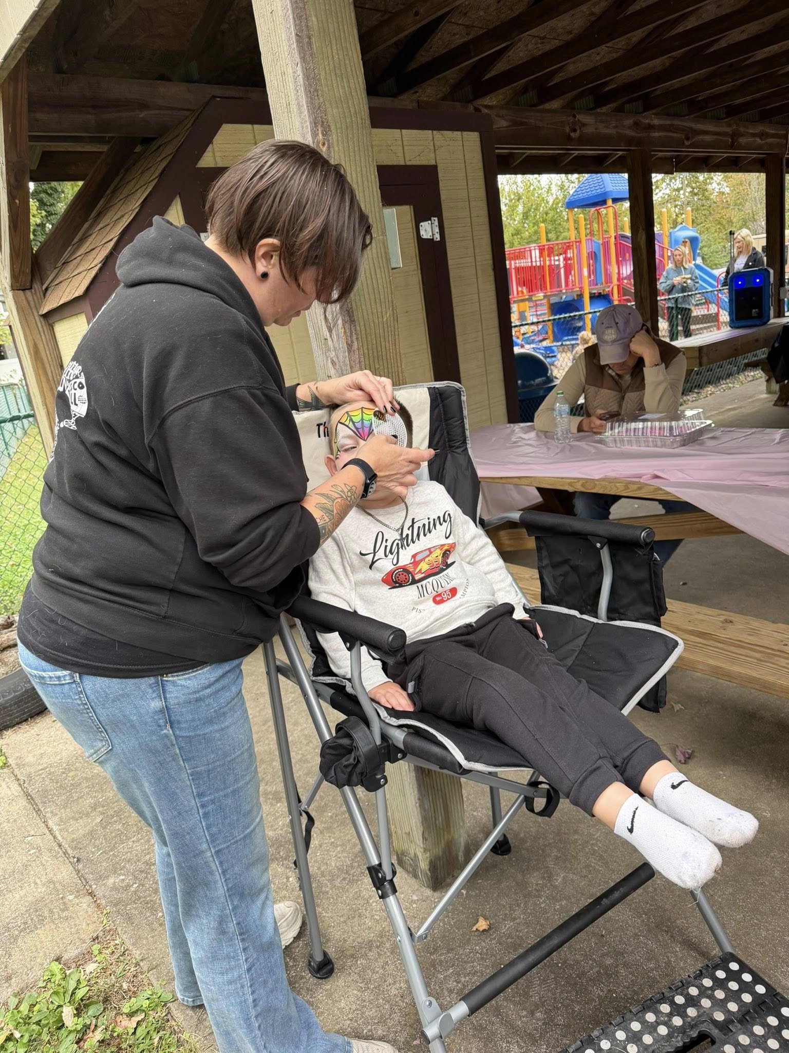 A woman doing face painting on a girl sitting in a stroller at an outdoor event near a playground.