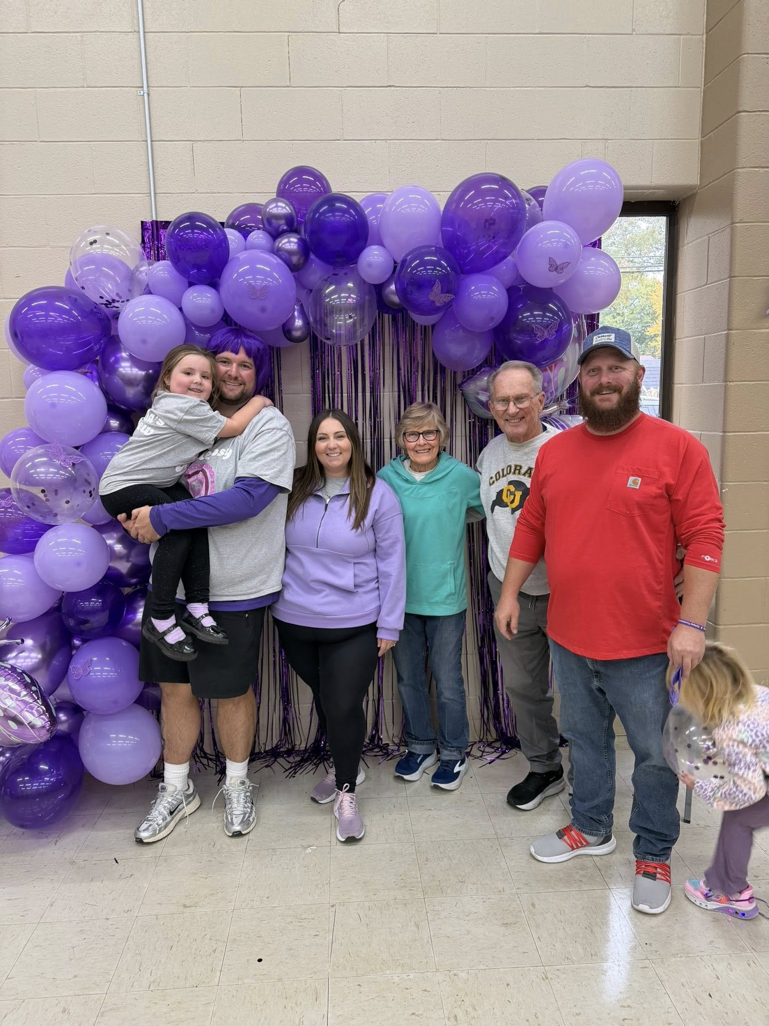 Group of six people standing in front of purple balloon arch with shiny and butterfly designs, indoors with beige walls and window.