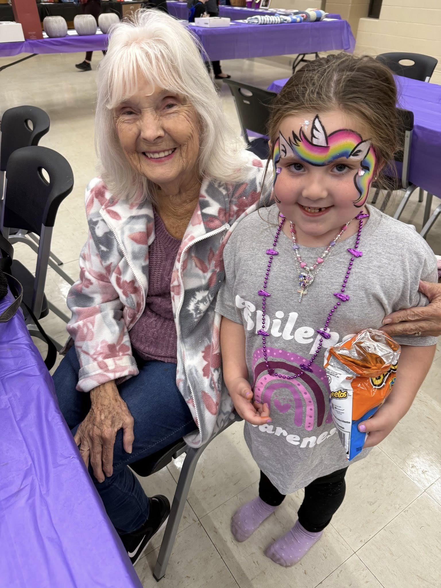 An elderly woman and a young girl are smiling at a party. The girl has face paint of a colorful rainbow unicorn on her face and is wearing necklaces and holding a bag of Cheetos. The woman is wearing a patterned fleece jacket and a purple top. They a