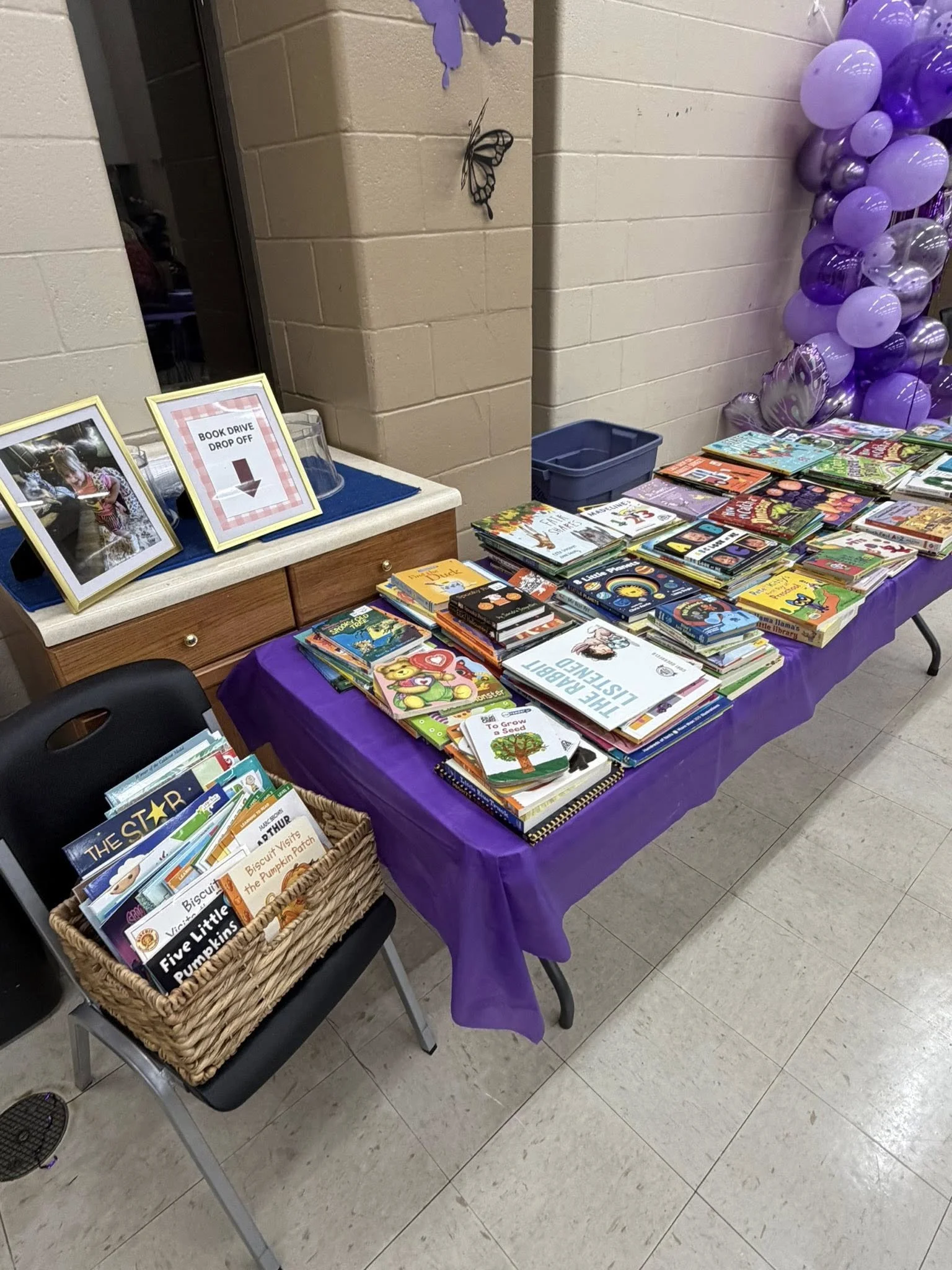 Table covered with purple cloth filled with children's books, set up for a book drive, with balloons on the side.